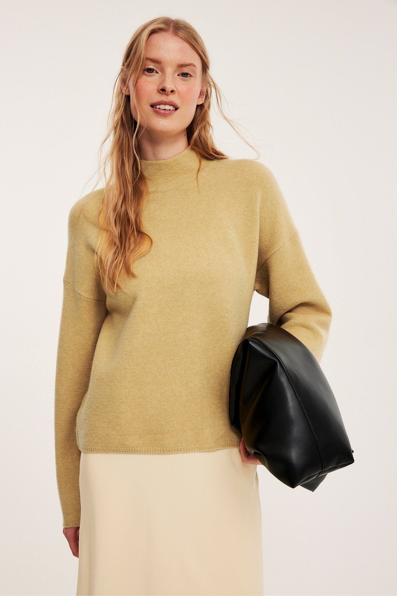 Frontbilde: Kvinne i lys gul strukturert turtleneck-genser, lys beige skjørt, holder en svart veske.