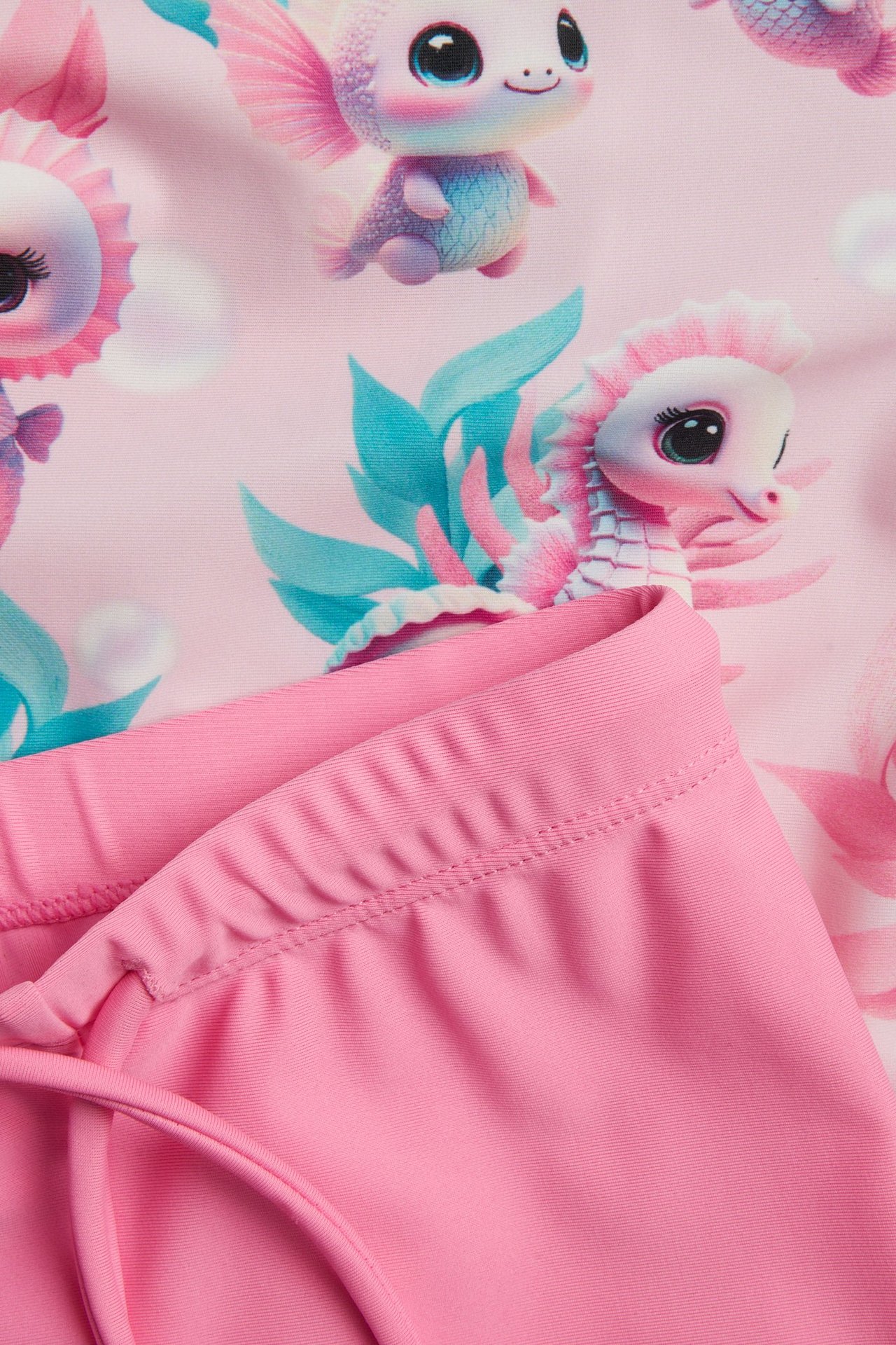 Lähikuva lasten vaaleanpunaisesta kuvioidusta UV-paidasta ja pinkeistä UV-housuista kiristysnyörillä.