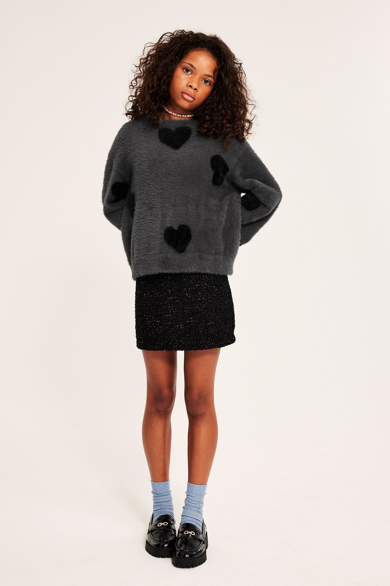 Framsida av ett barn i mörkgrå fluffig tröja med svarta hjärtan, svart kjol, blå strumpor och svarta loafers.
