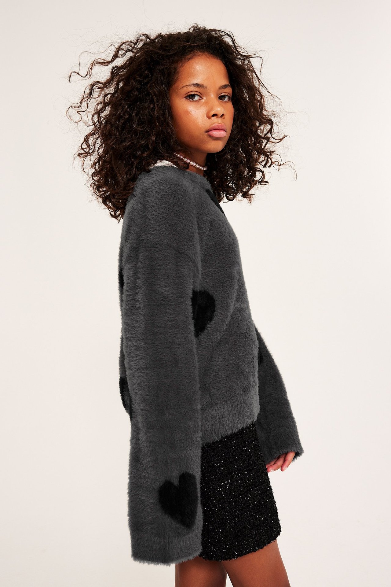 Sidovy: Ett barn bär en mjuk grå fjädergarnströja med svarta jacquardhjärtan och en svart glittrig kjol.