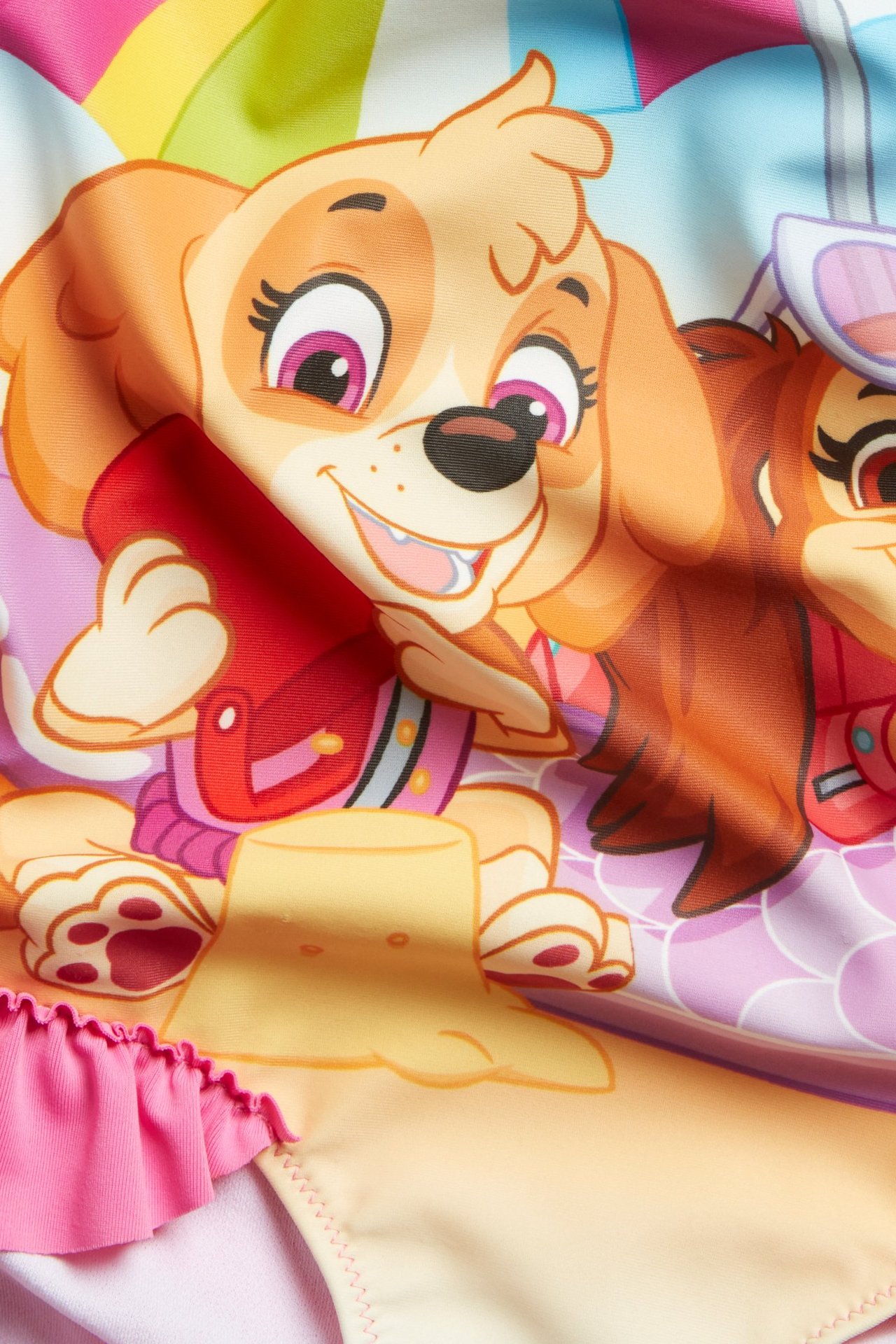 Lähikuva vaaleanpunaisesta lasten uima-asusta, jossa on Ryhmä Hau -printti edessä ja röyhelö.