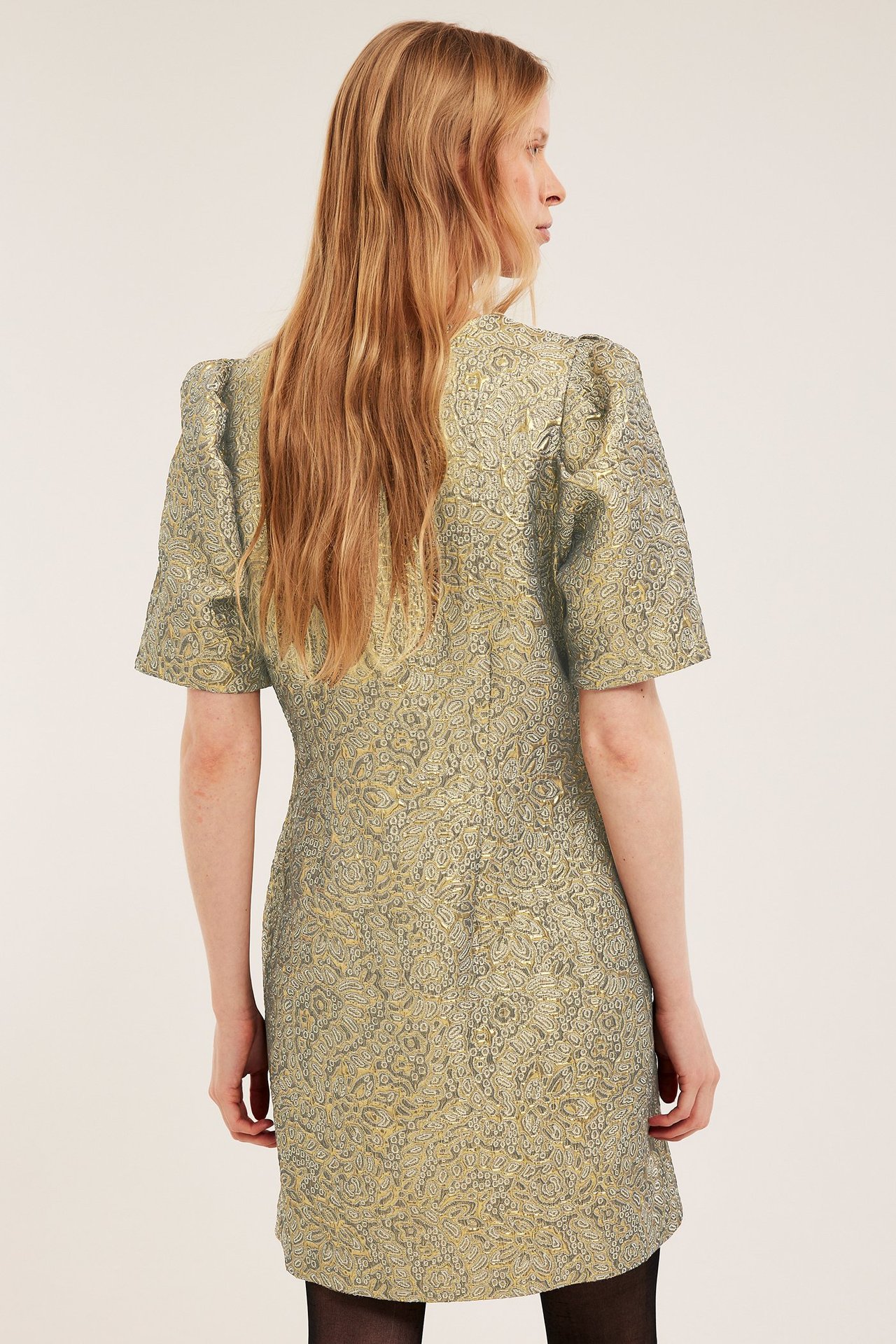 Bakifrån: kvinna i guldfärgad jacquardbrokadklänning med korta puffärmar.