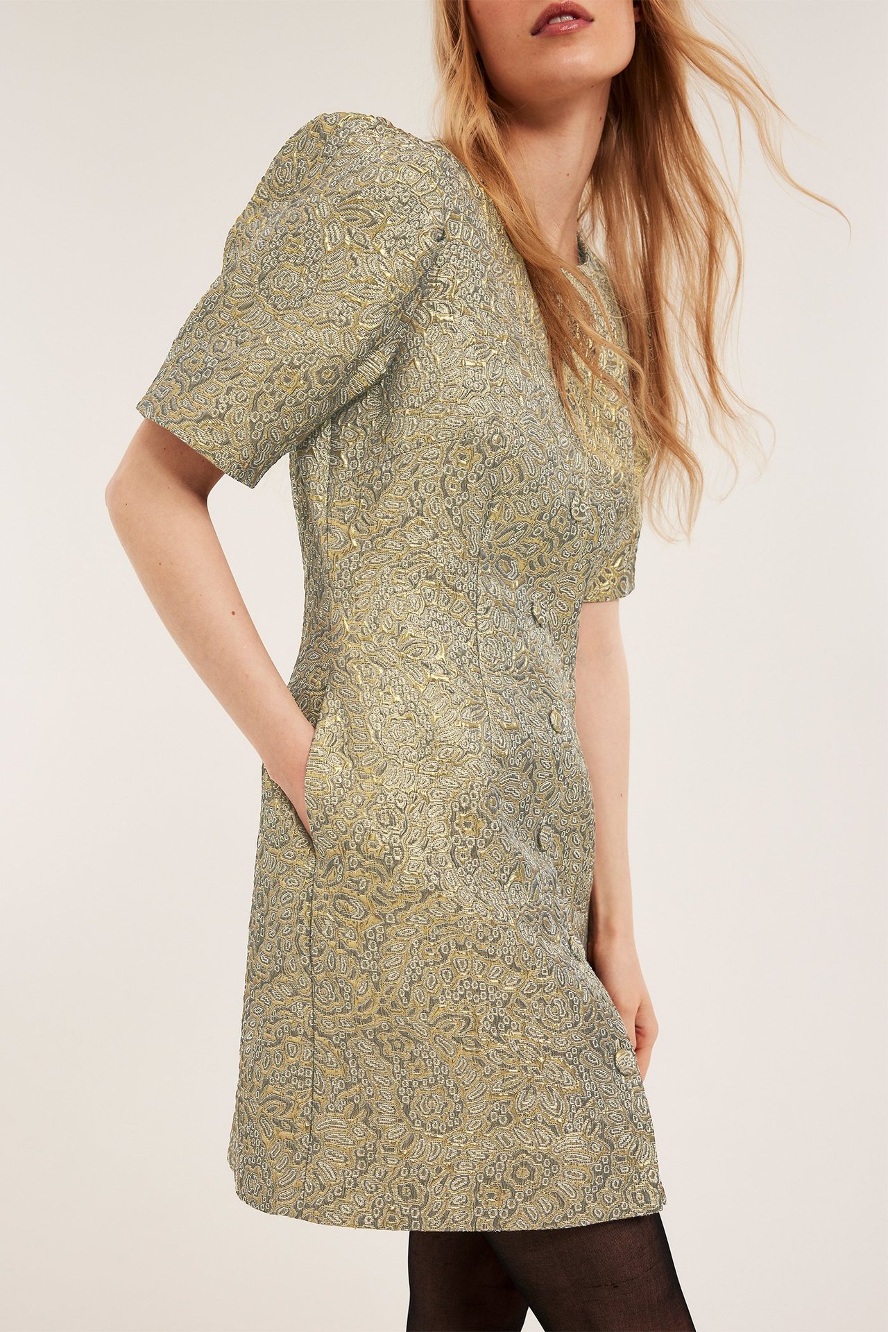 Sned framifrån: Kvinna i guldfärgad jacquardbrokadklänning med korta puffärmar, fickor och svarta strumpbyxor.