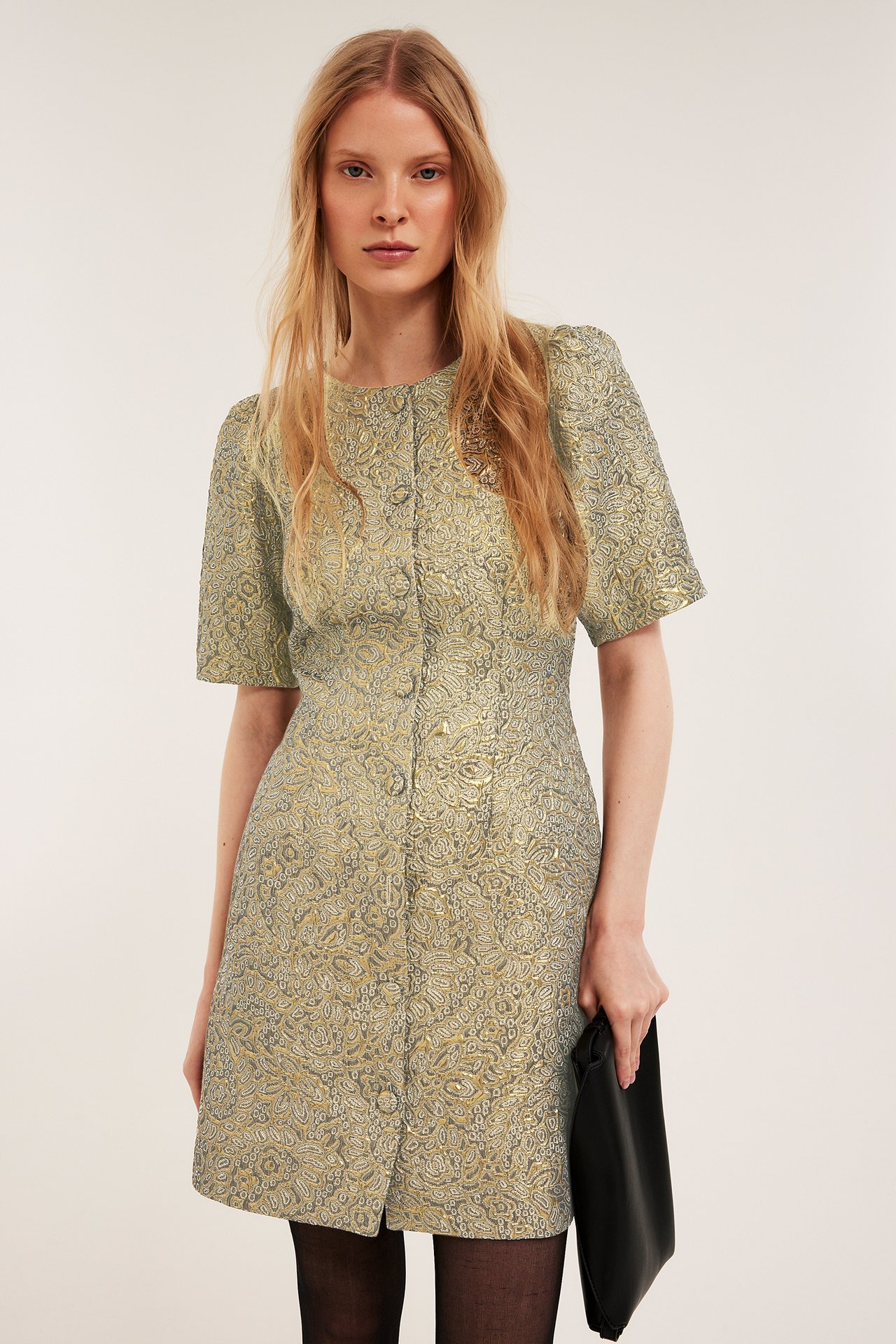 Framifrån: Kvinna i guldbrokad kortärmad klänning med knappar, håller svart clutchväska och svarta strumpbyxor.