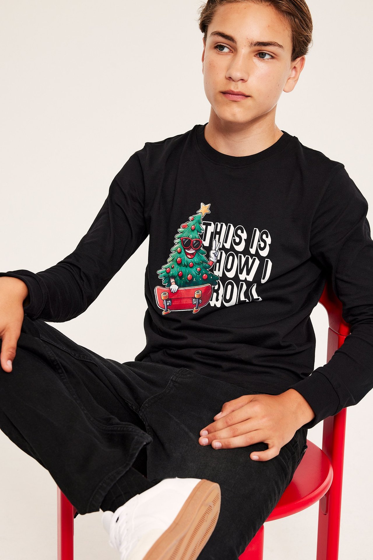 Z przodu: dziecko w czarnym t-shircie z długim rękawem z nadrukiem świątecznym, czarnych dżinsach i białych trampkach.