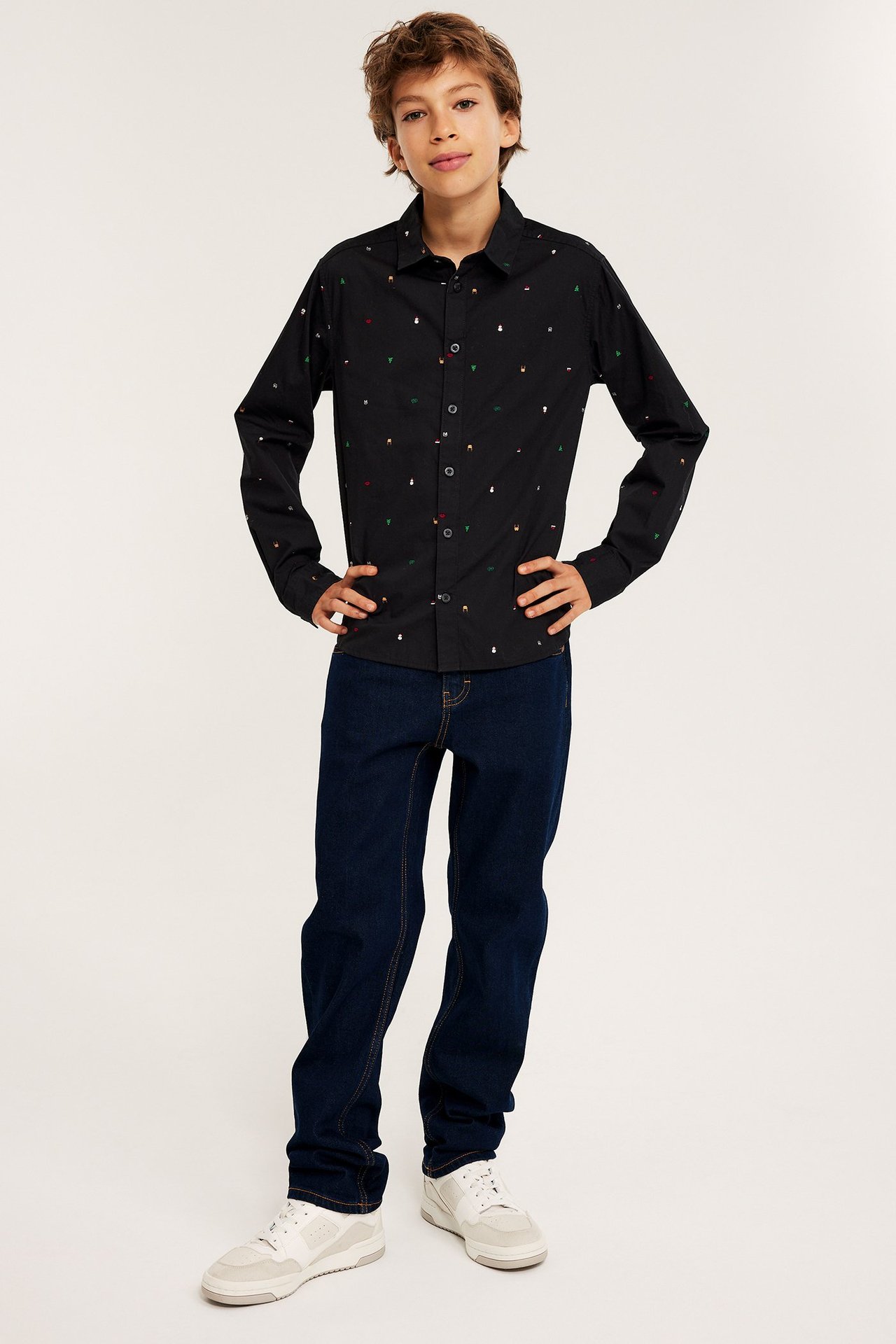 Edestäpäin, lapsi mustassa puuvillapaidassa värikkäillä jouluaiheilla, tummansiniset farkut ja valkoiset lenkkarit.