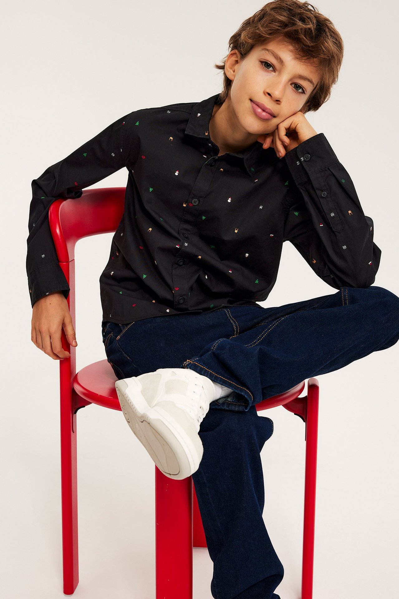 Widok z przodu dziecka w czarnej bawełnianej koszuli ze świątecznym wzorem, ciemnoniebieskich dżinsach i białych trampkach.