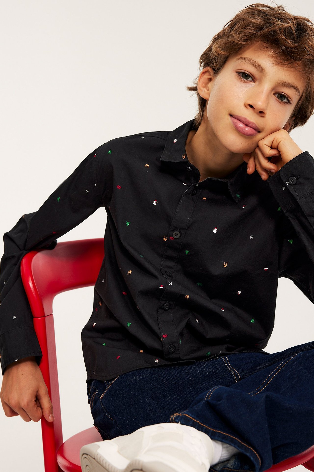 Widok z przodu: dziecko w czarnej bawełnianej koszuli z kolorowymi świątecznymi wzorami, ciemnoniebieskich jeansach i białych sneakersach.