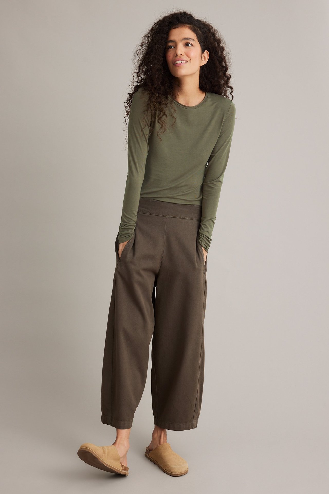 Frontbilde: Kvinne i en khakigrønn langermet topp, mørkebrune vide bukser og lysebrune tresko.
