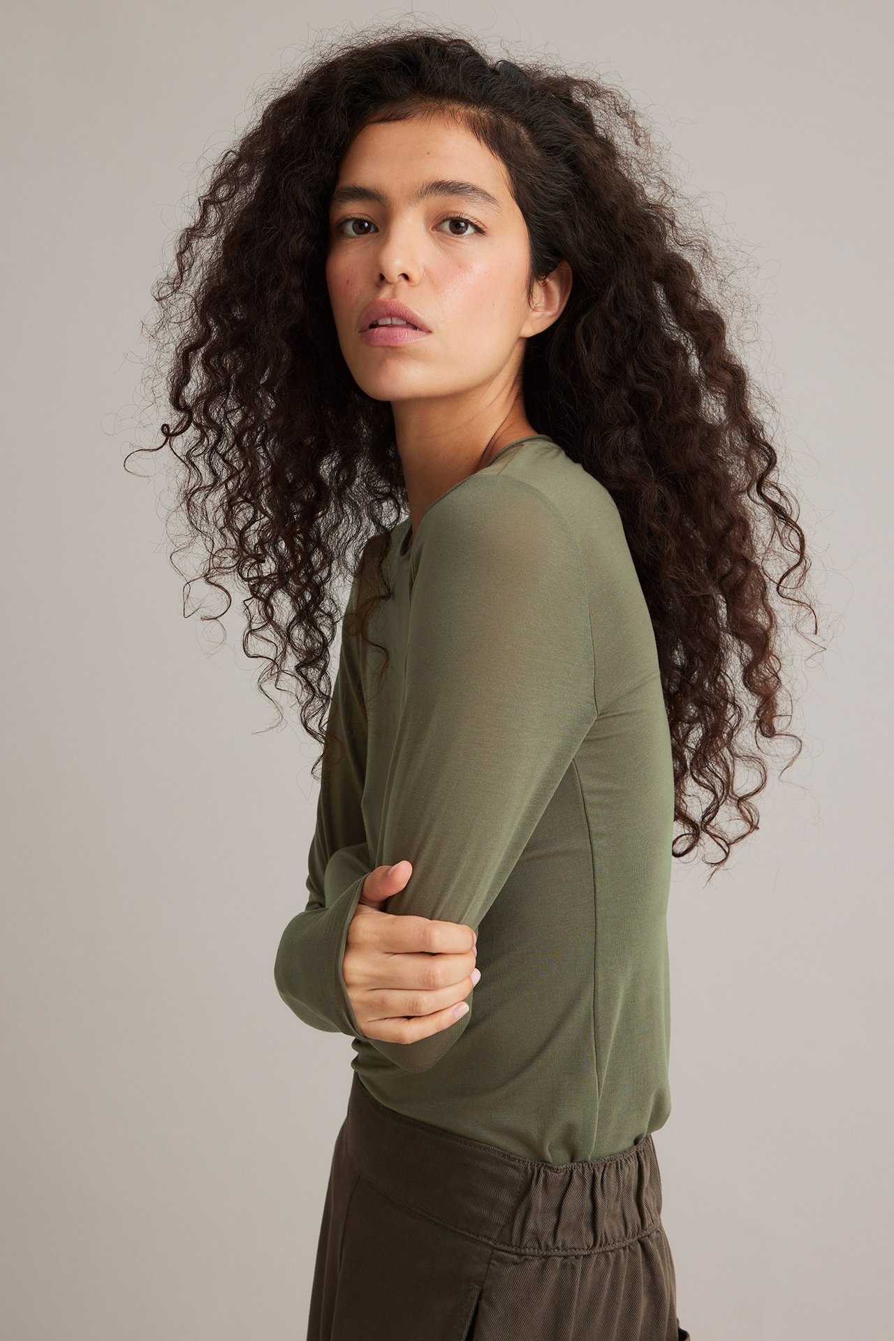 Sidebilde av kvinne i en khakigrønn rundhalset langermet modaltop og mørkebrune bukser.