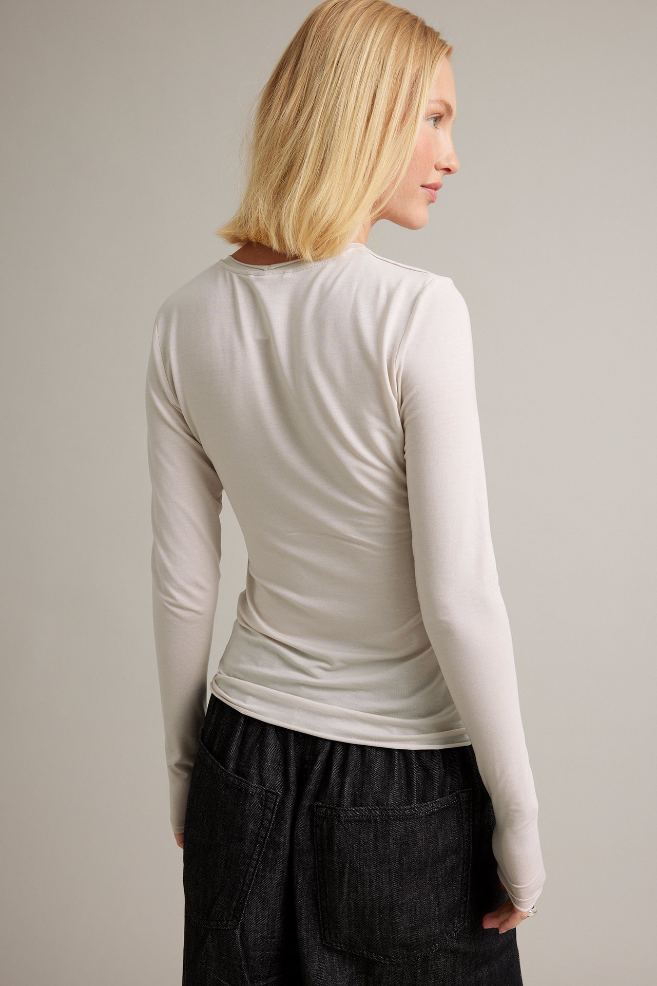 Bakfra av kvinnelig modell i en lys beige langermet topp og mørke vide jeans.