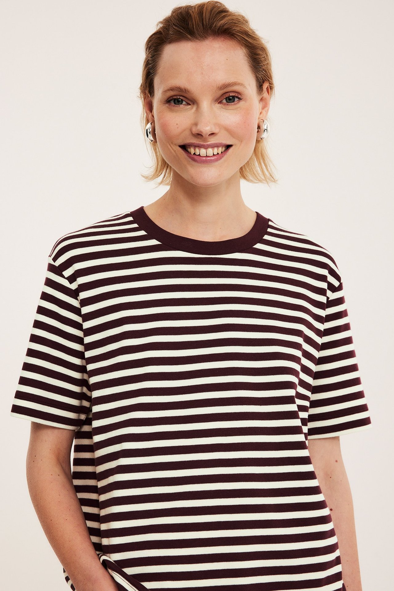 Nærbilde forfra av en kvinne i en oversized offwhite og mørkerød stripete bomulls-T-skjorte og sølvfargede øreringer.