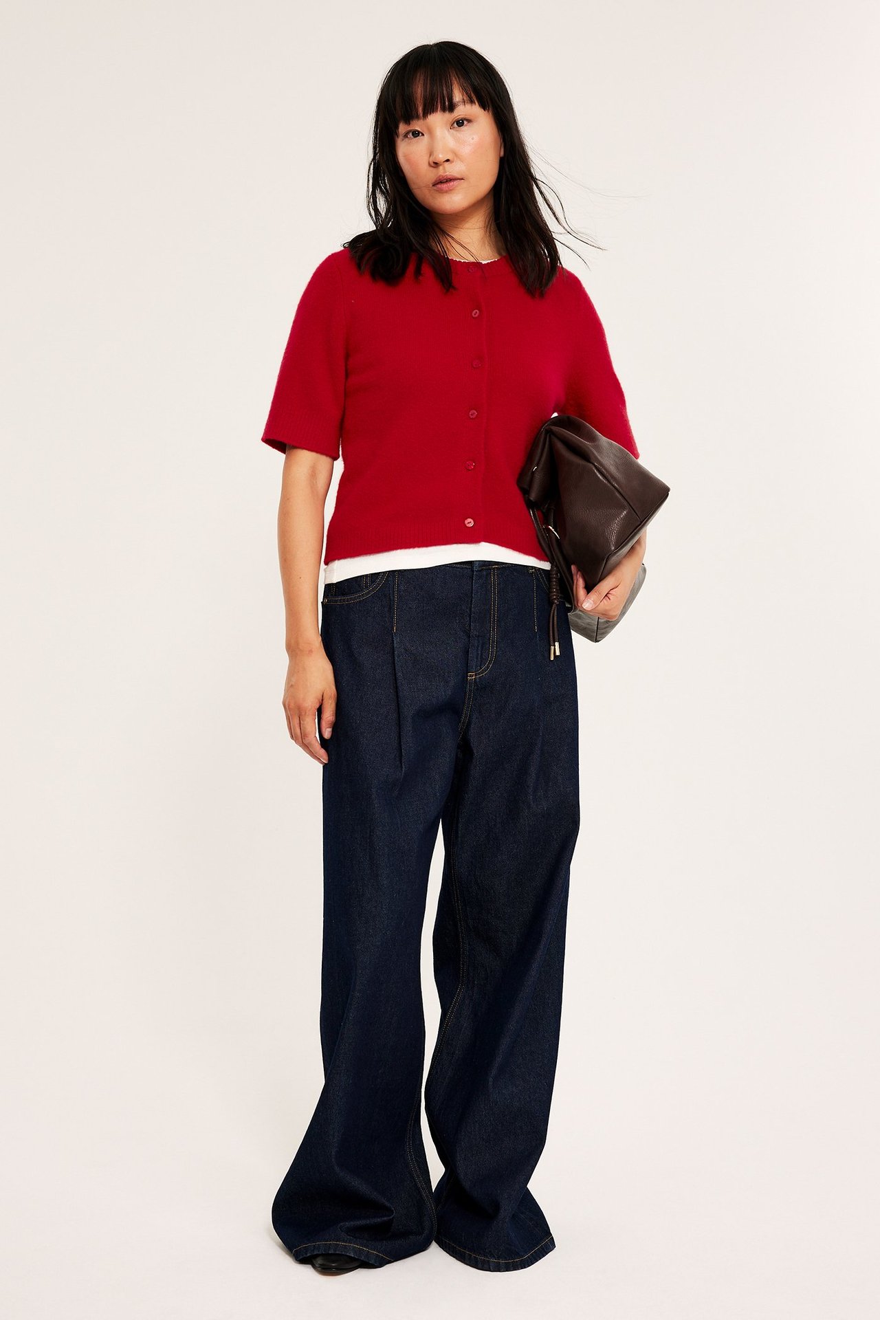 Kvinne i rød kortermet cardigan, mørke jeans, holder brun veske, front.