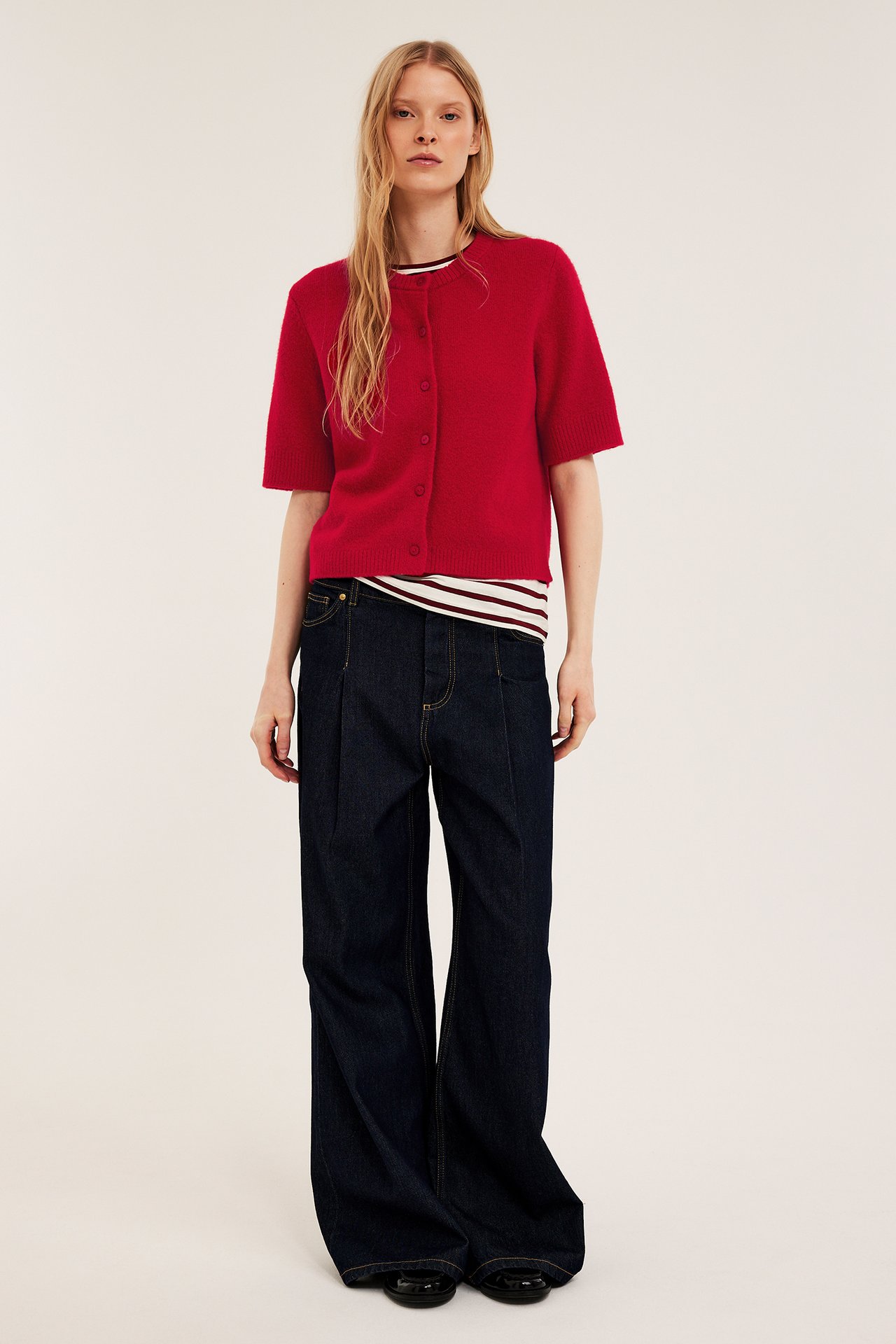 Framifrån: Kvinna i röd kortärmad stickad kofta med knappar, randig topp, mörkblå vida jeans och svarta skor.