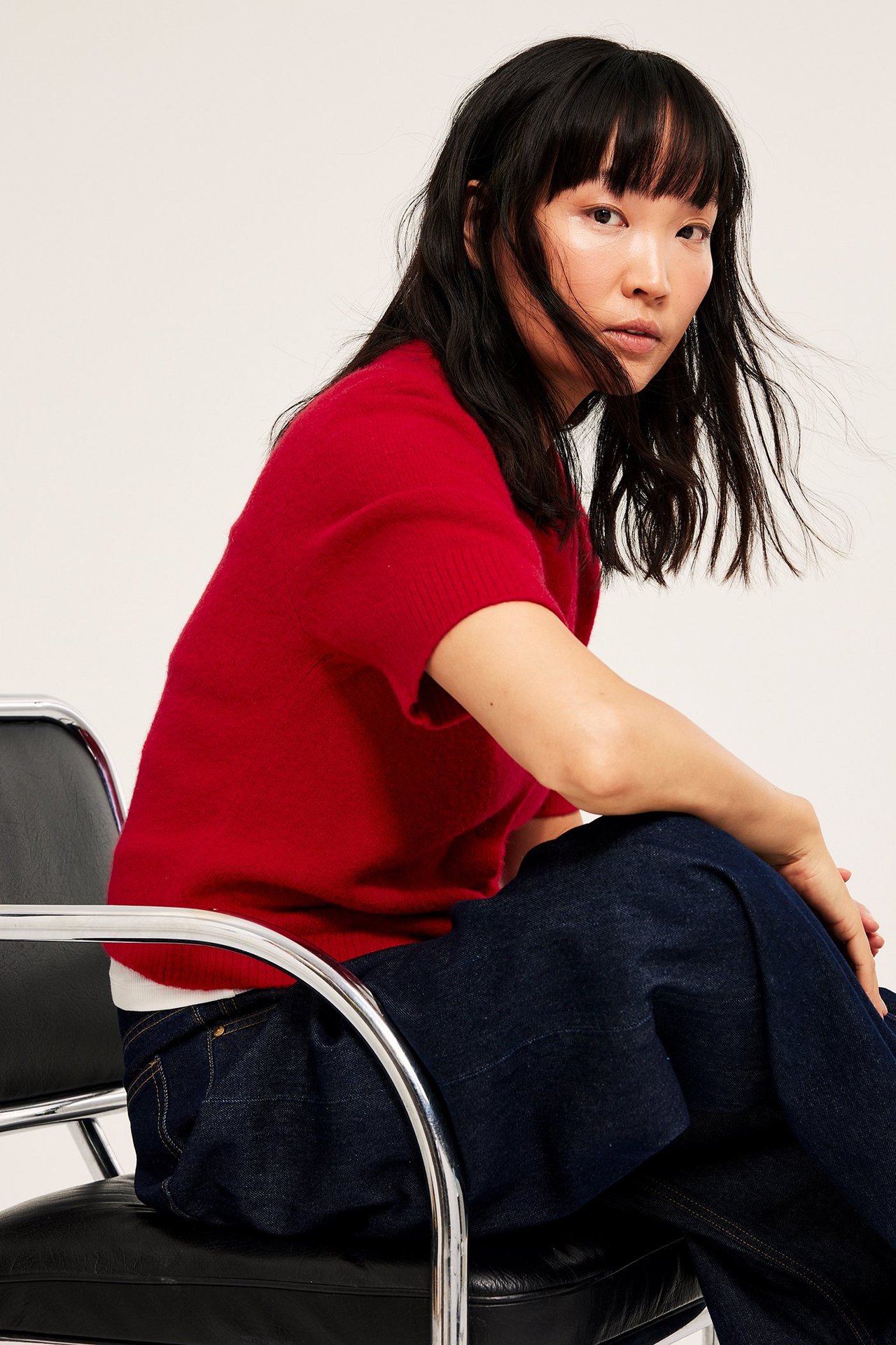 Kvinne i rød kortermet cardigan, sitter, svart jeans, enkel bakgrunn.