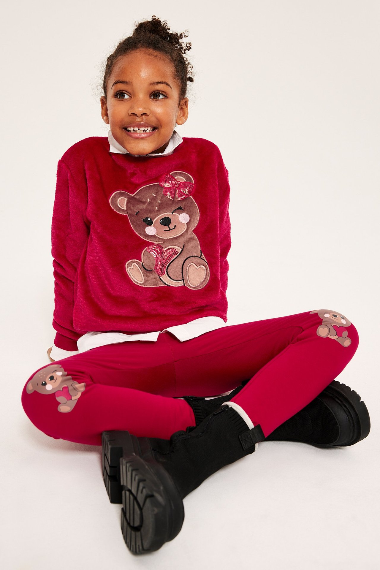 Dziewczynka w czerwonych legginsach z aplikacją misia na kolanach, siedzi na białym tle.