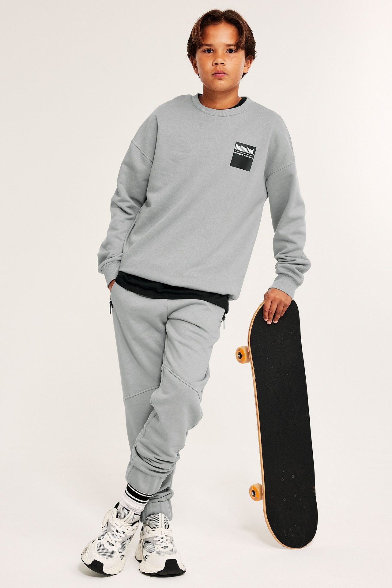 Chłopiec w szarej bluzie z nadrukiem na piersi, trzyma deskorolkę, ściągacze i wykończenie.