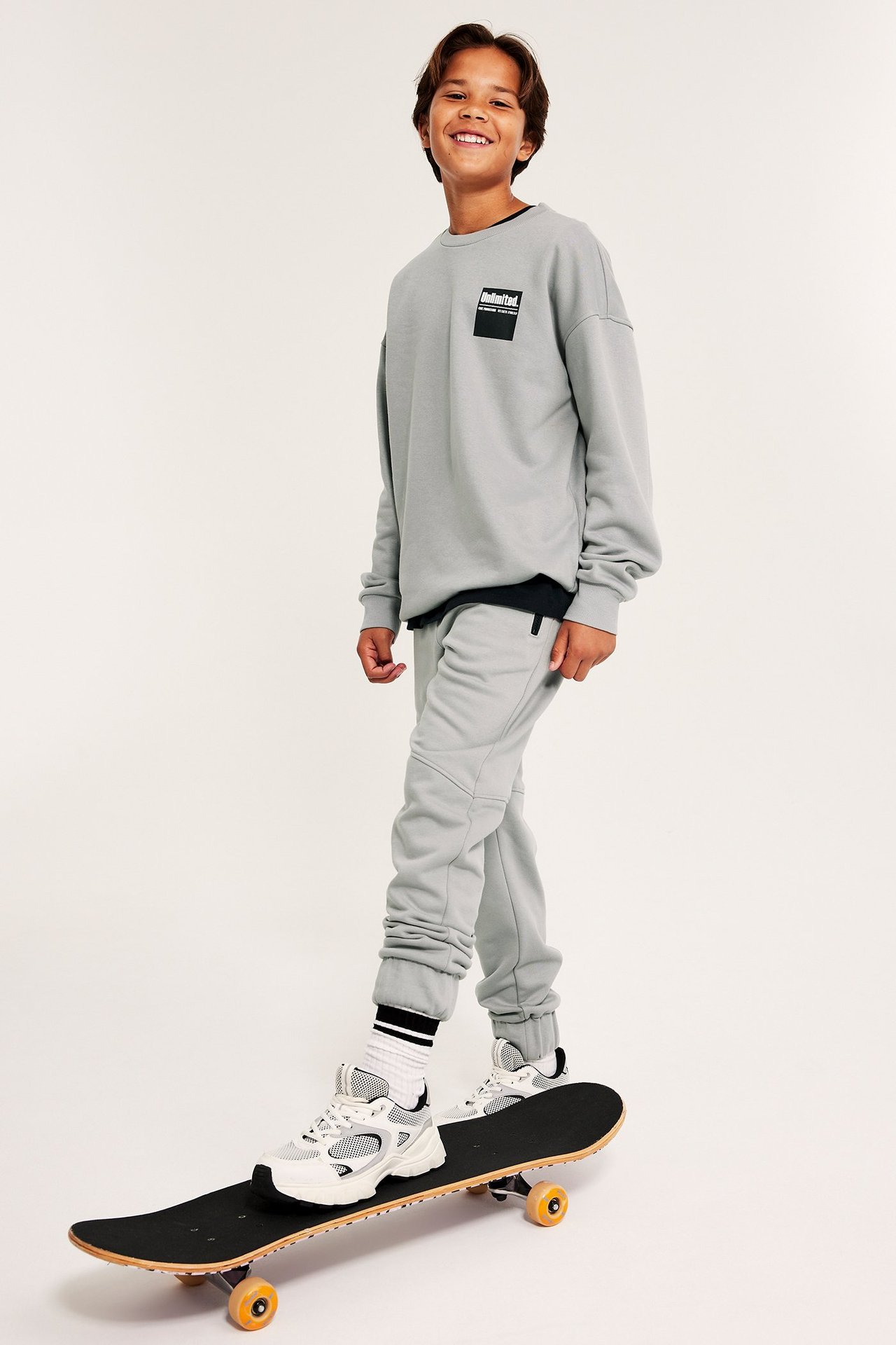 Chłopak w szarych spodniach i bluzie, stoi na deskorolce, widok z przodu.