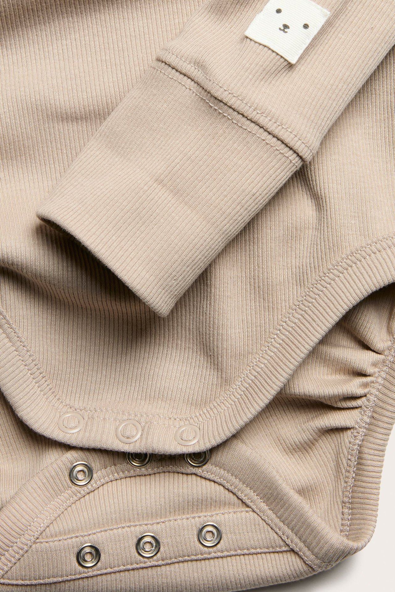 Närbild på en beige ribbad babybody med långa ärmar, vikbara muddar, tryckknappar och en björnetikett.