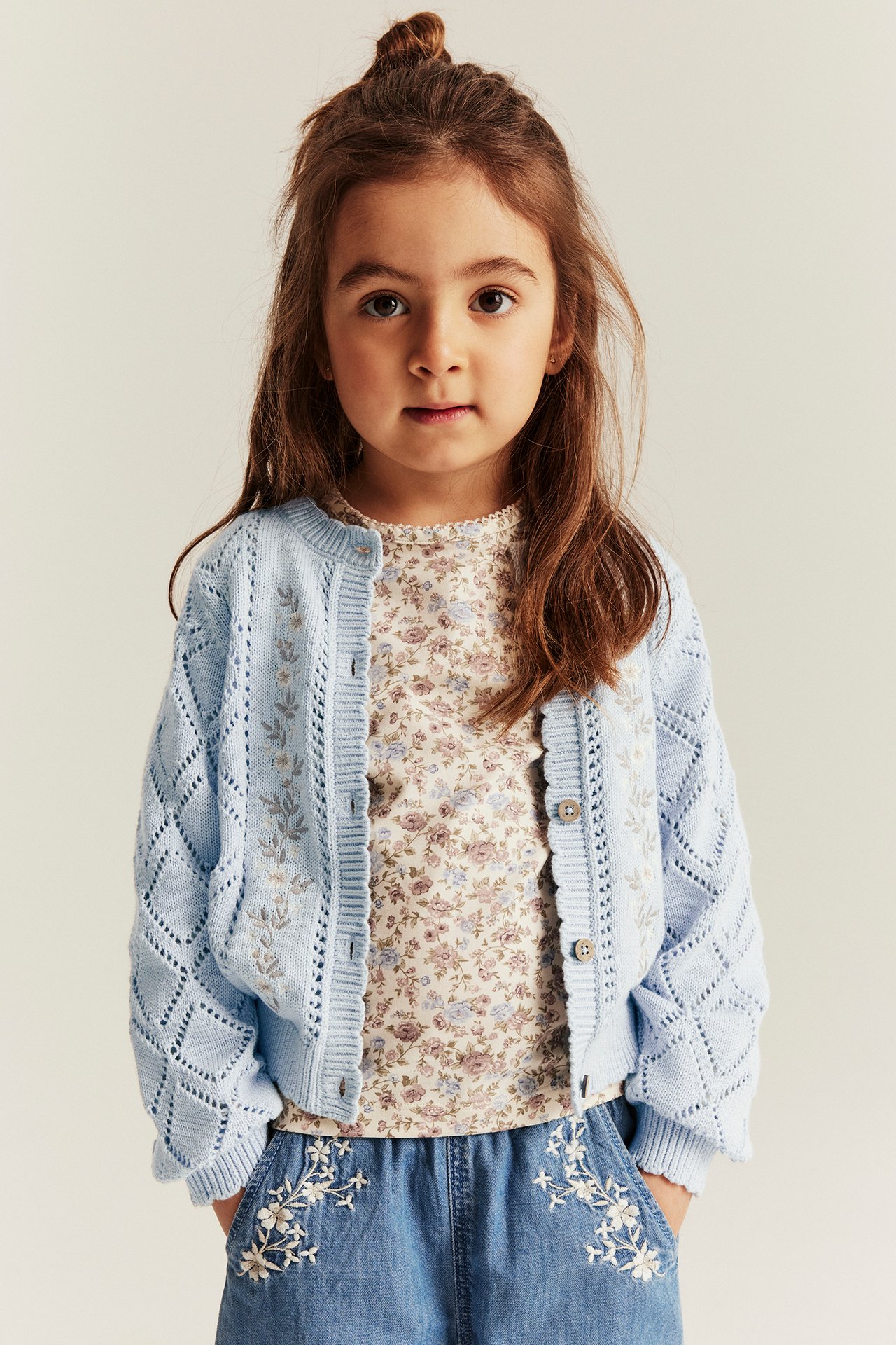 Edestä: Lapsi vaaleansinisessä kirjotussa neuletakissa, kermanvärinen kukkatoppi ja siniset kirjotut shortsit.