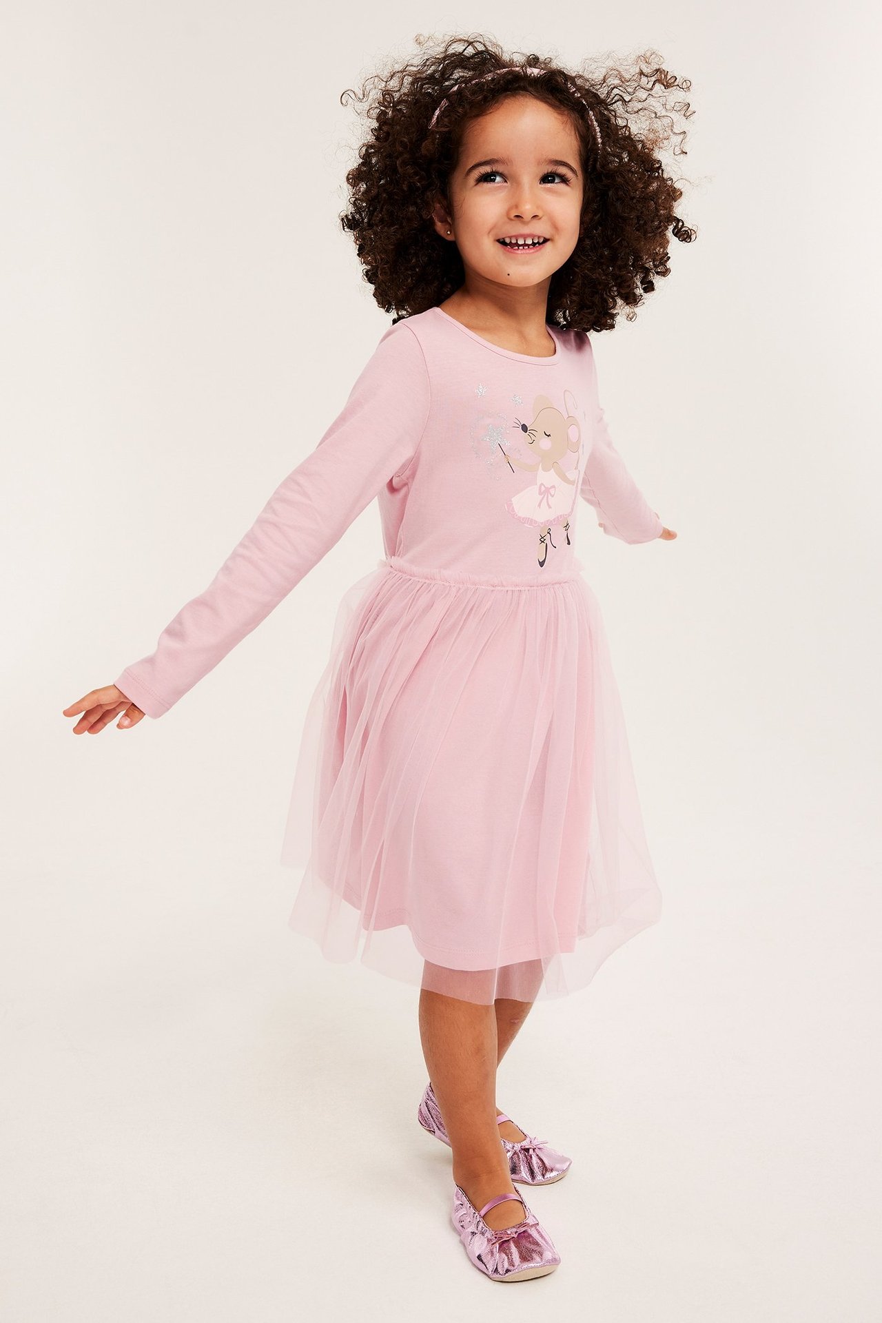 Edestä: Hymyilevä lapsi yllään vaaleanpunainen pitkähihainen mekko hiiripainatuksella, tyllihame ja metalliset pinkit kengät.