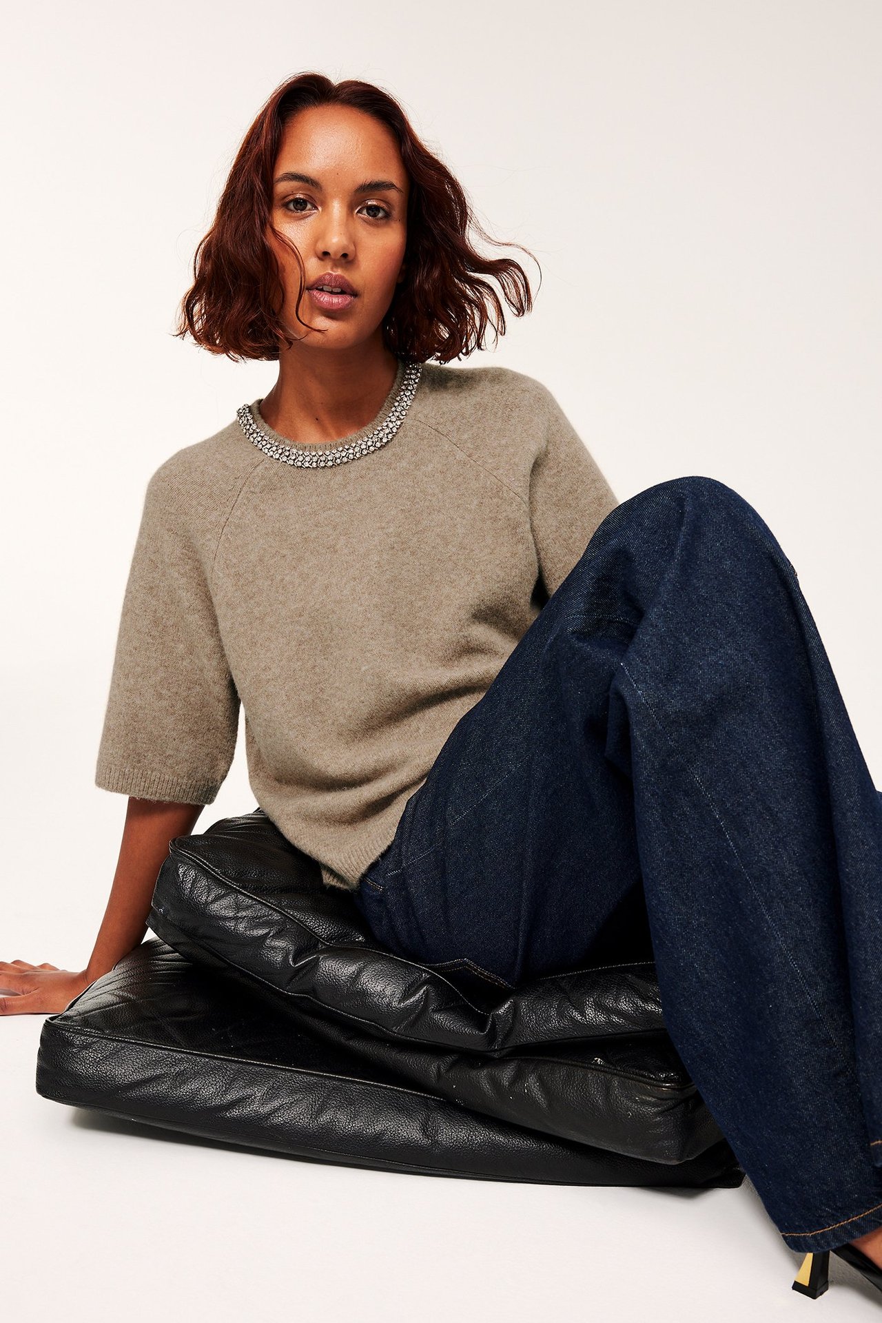 Kvinna i beige stickad tröja med halssmycke, mörkblå jeans, sittande på svart sittsäck.
