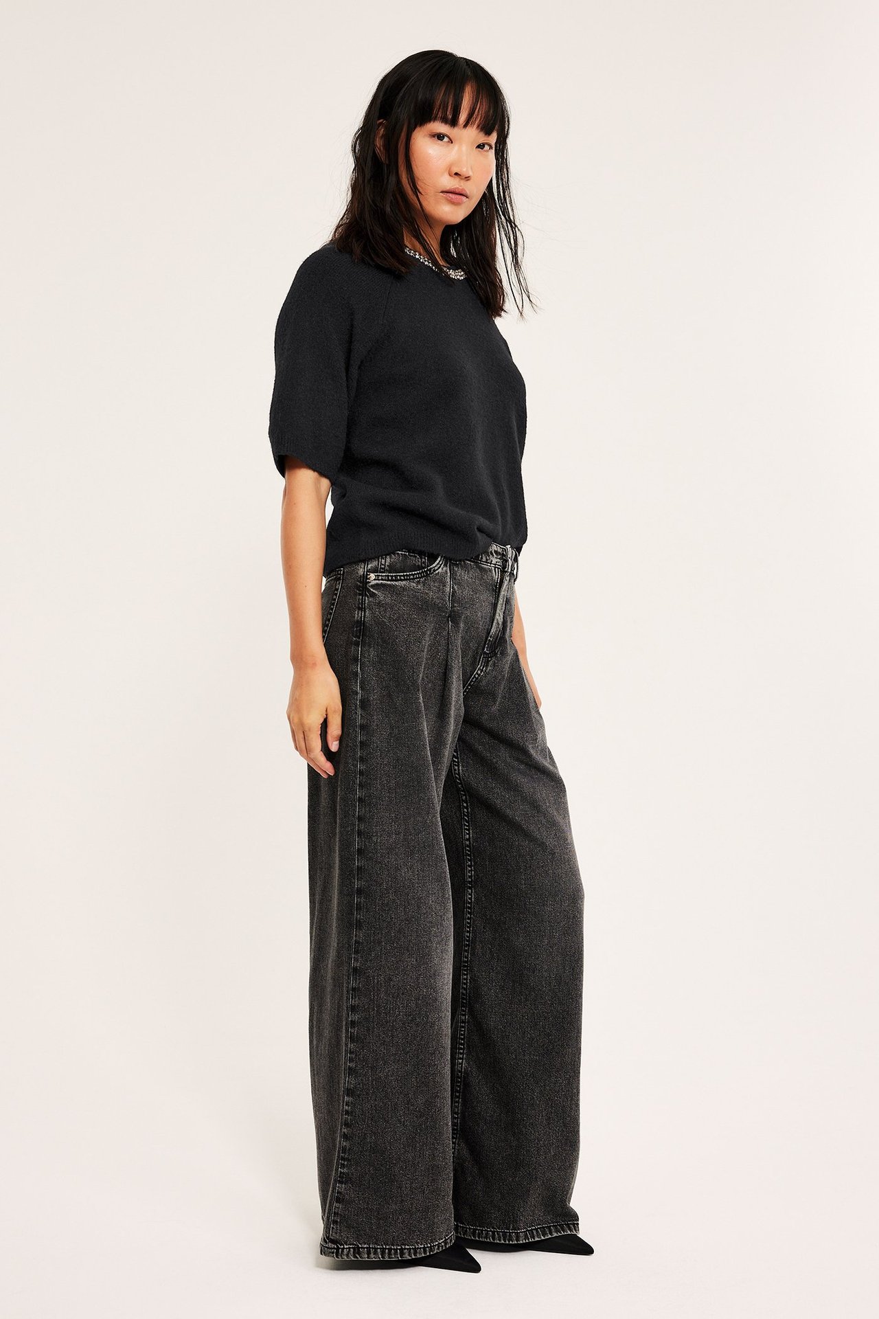 Svart stickad tröja med kort ärm, halsbanddetaljer, över svart vida jeans, frontal vy.