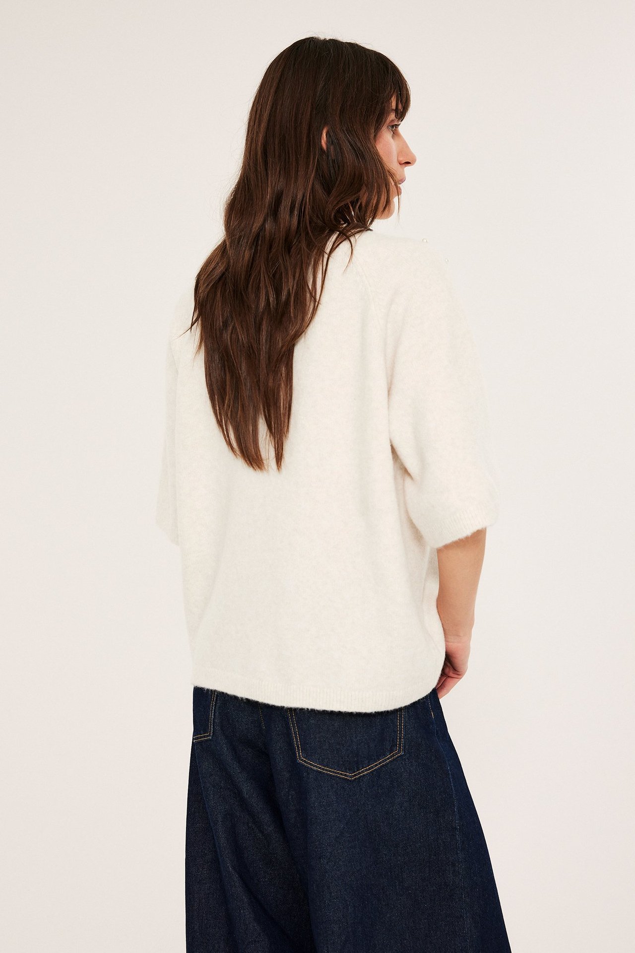 Kvinna i vit stickad tröja med kort ärm, halsband, pärlor, framför, mörkblå jeans.