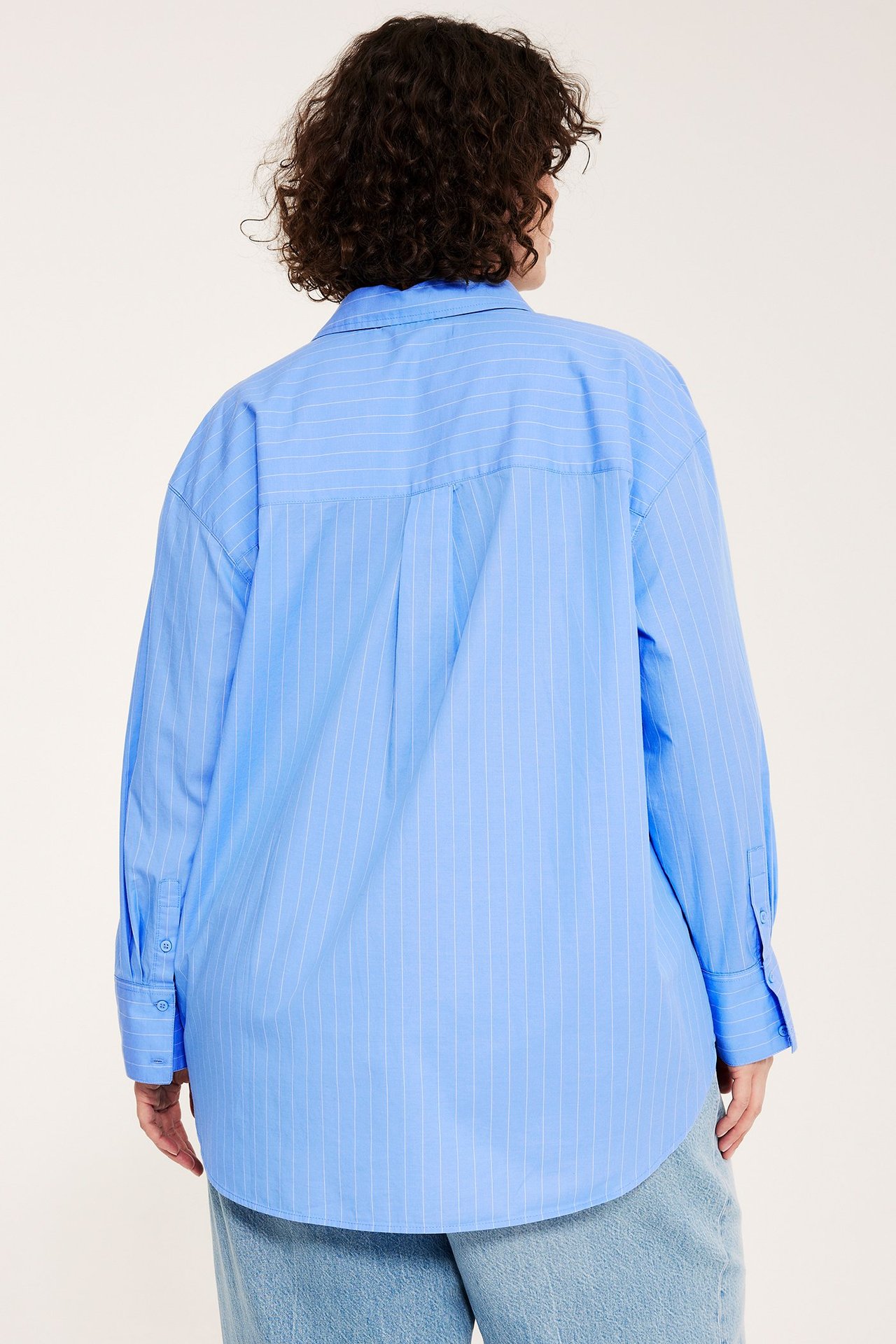 Bakifrån: kvinna i oversized ljusblå randig bomullsskjorta och ljusblå jeans.