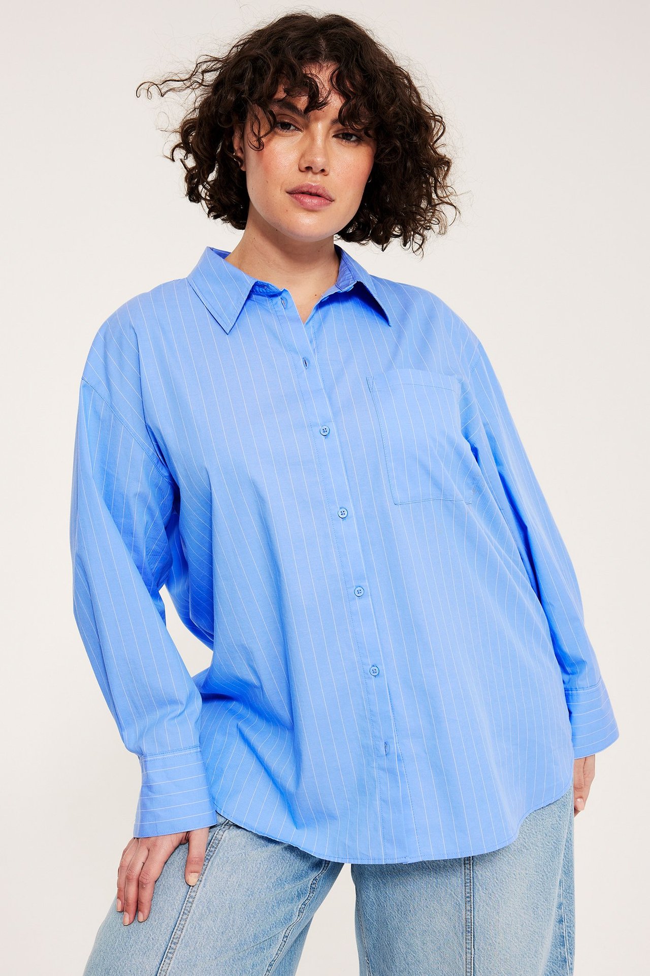 Edestäpäin kuvattu nainen yllään oversized sininen raidallinen puuvillapopliinipaita rintataskulla, vaaleiden farkkujen päällä.