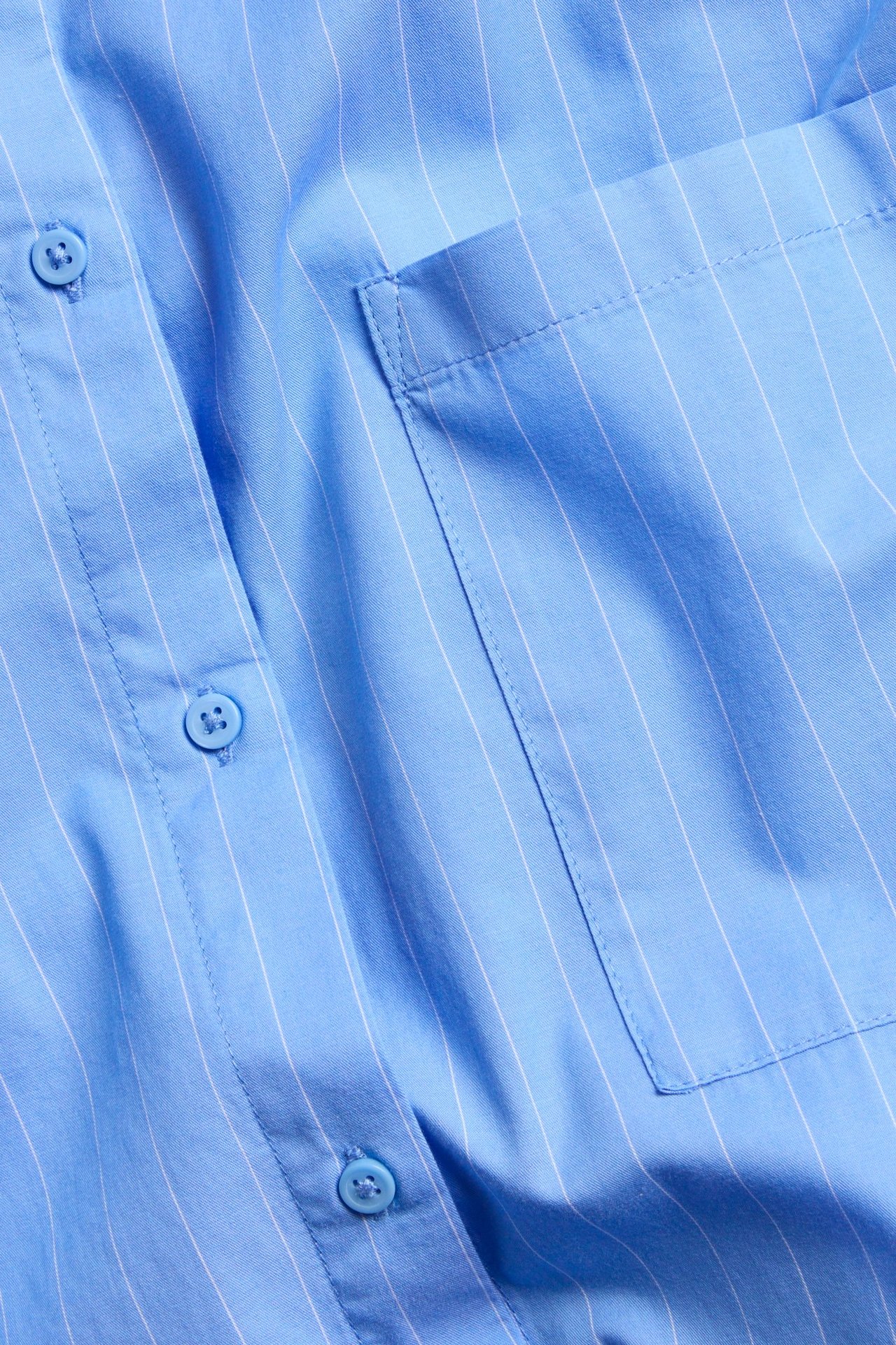 Lähikuva vaaleansinisestä raidallisesta popliinipaidasta, jossa on valkoisia raitoja, sinisiä nappeja ja rintatasku.