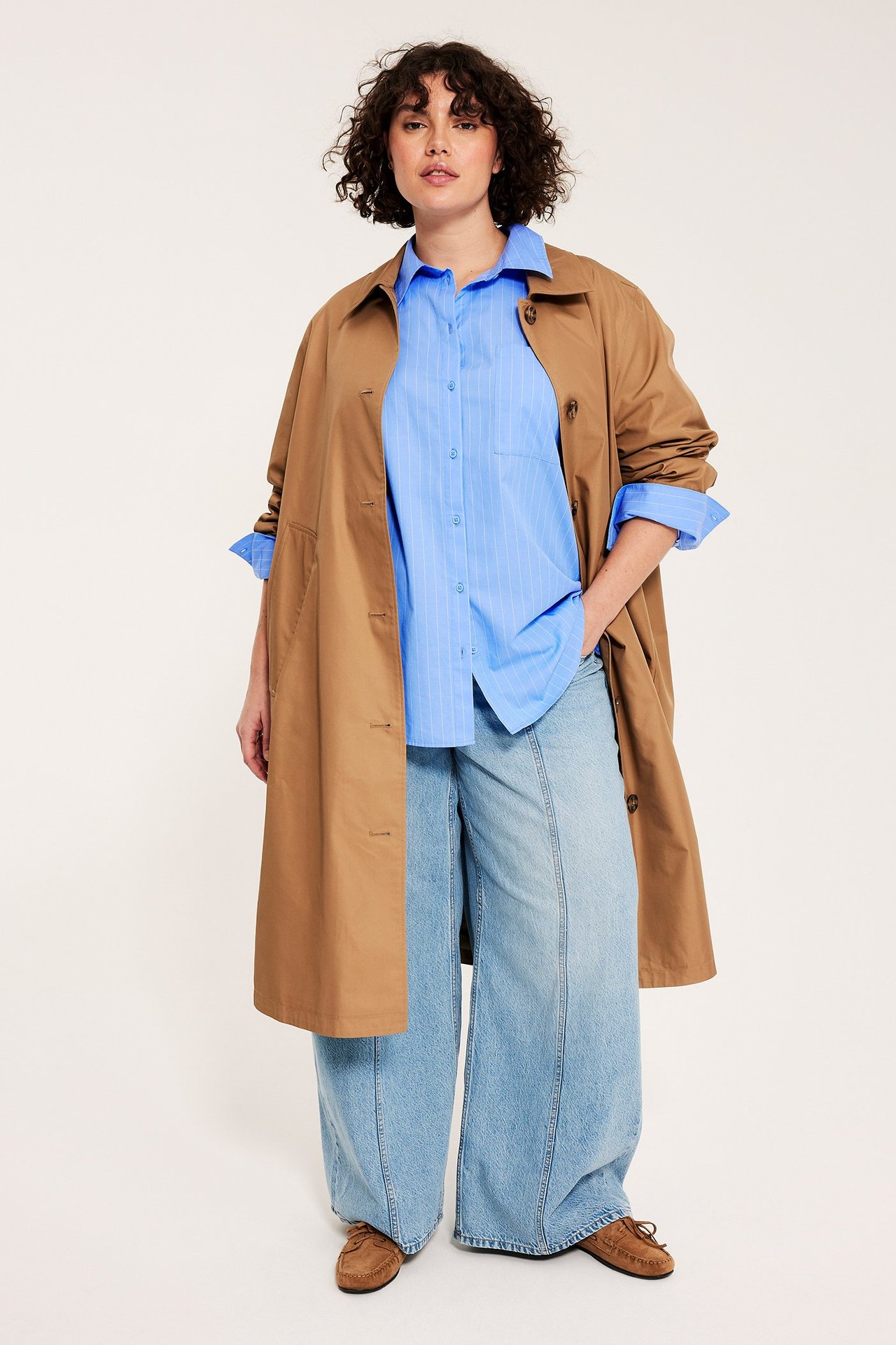 Nainen edestä: siniraitainen popliinipaita, ruskea trenssitakki, vaaleansiniset farkut ja mokkasiinit.