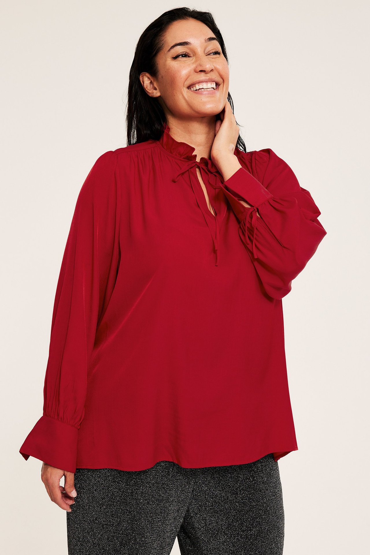 Widok z przodu kobiety w czerwonej bluzce z długim rękawem z falbanką i wiązaniami, na ciemnych spodniach.