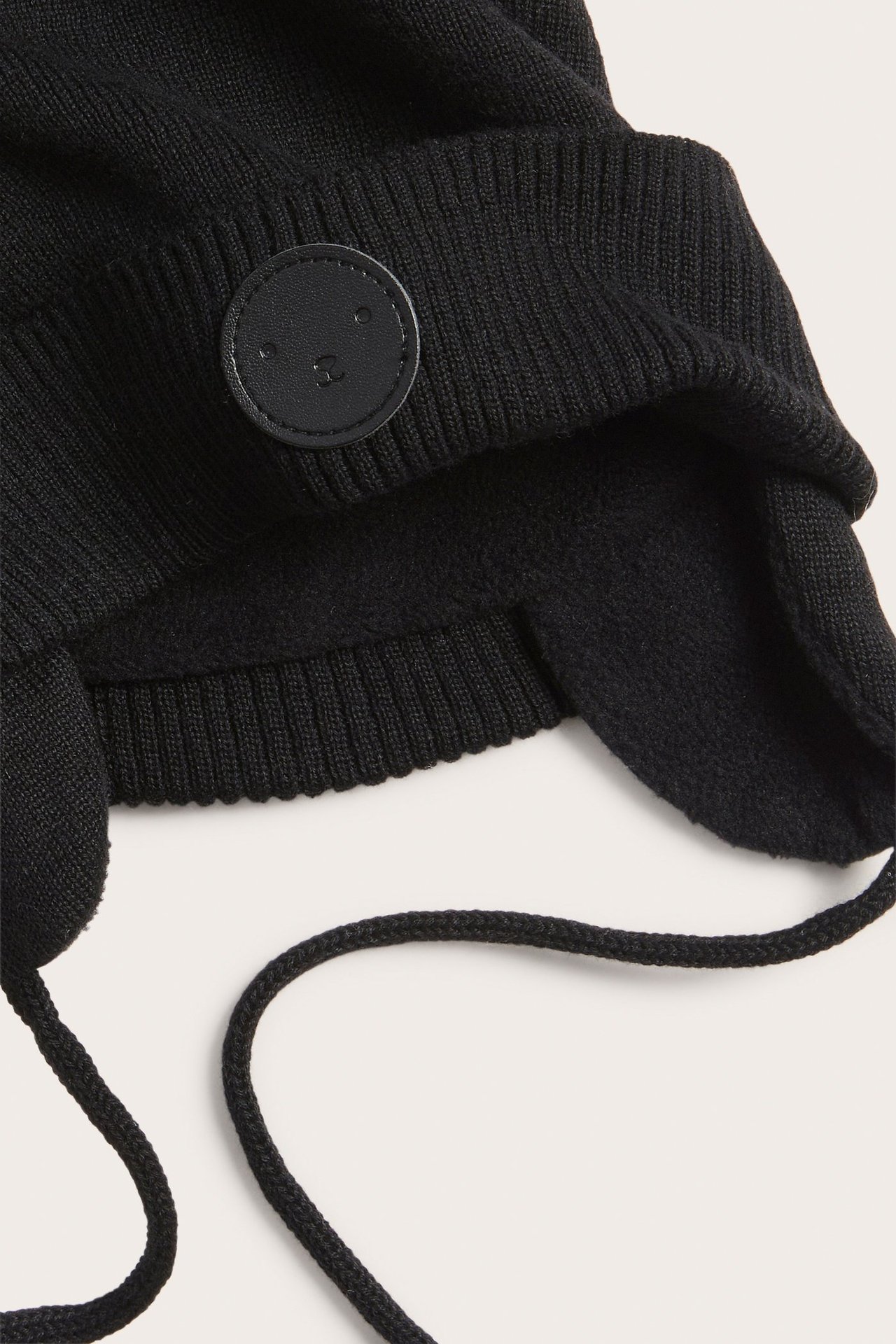 Närbild av en svart stickad babymössa med öronlappar, knytband, fleecefoder och en liten etikett.