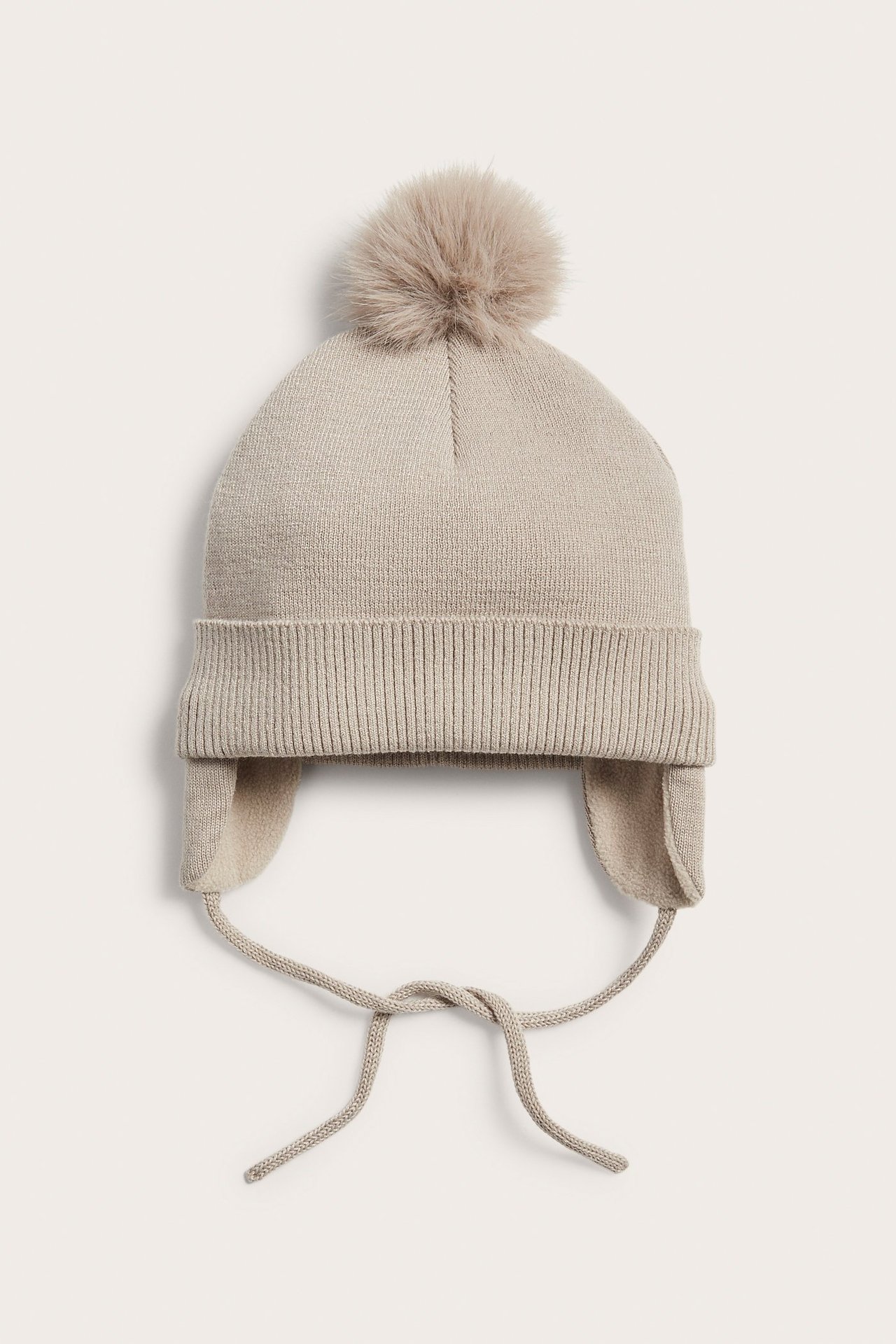 Framifrån: beige stickad babymössa med fuskpälsboll, öronlappar och knytband.