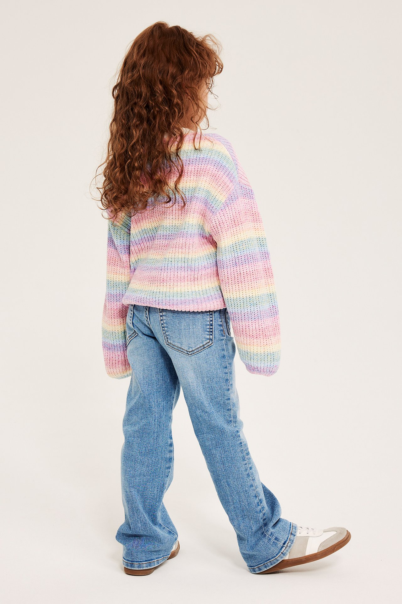 Widok z tyłu dziecka w jasnoniebieskich jeansach bootcut, pastelowym swetrze w tęczowe paski i białych sneakersach.