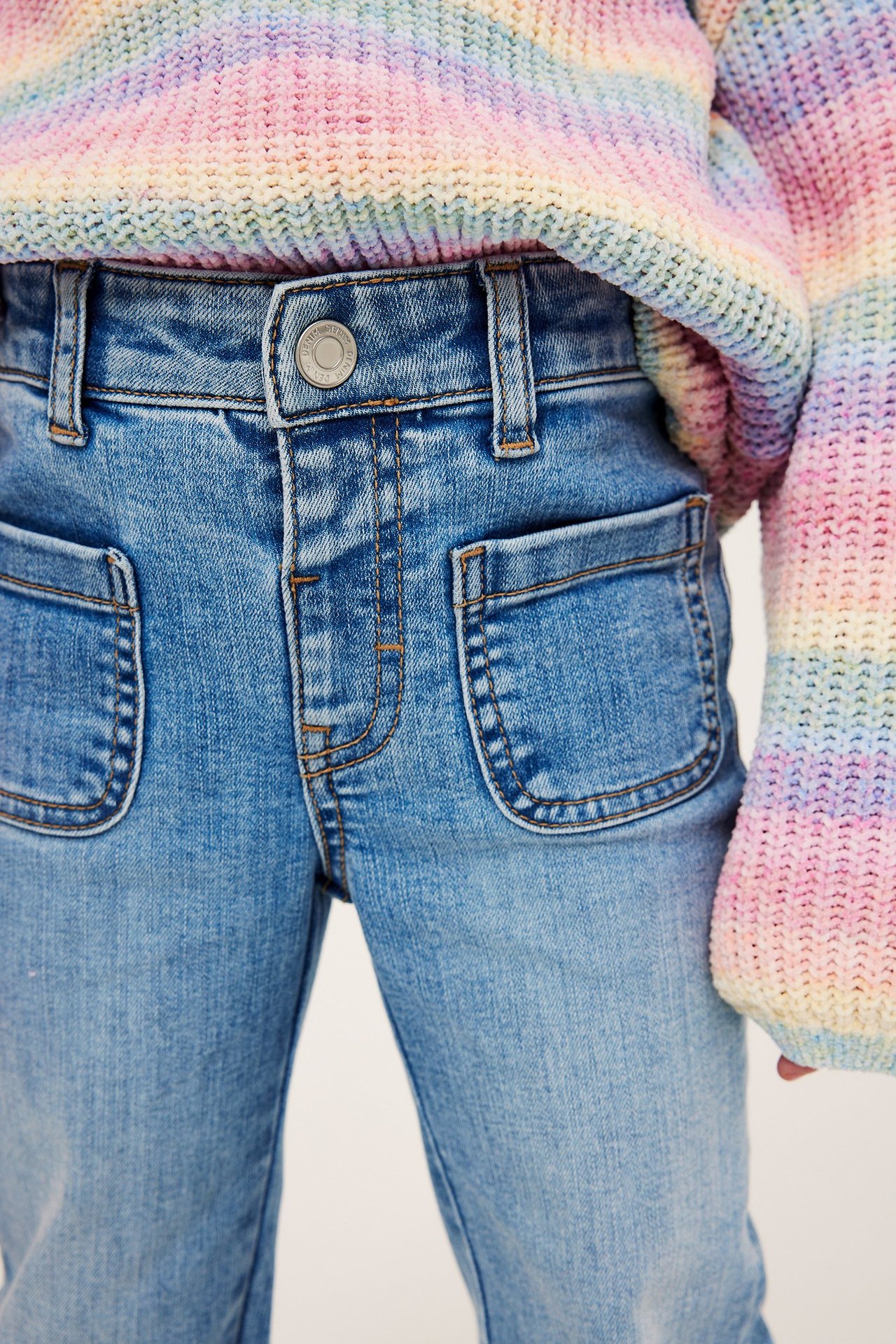 Lähikuva edestä lapsesta yllään vaaleansiniset joustavat bootcut-farkut retrotaskuilla ja sateenkaariraitaneule.