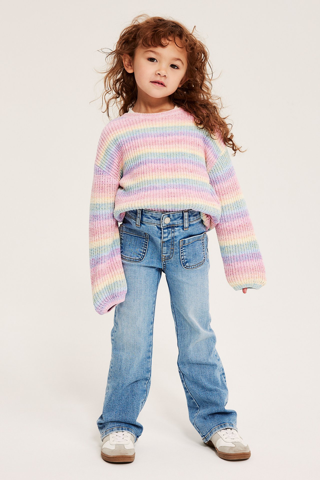 Widok z przodu dziecka w jasnoniebieskich dżinsach bootcut z naszywanymi kieszeniami i tęczowym swetrze w paski.