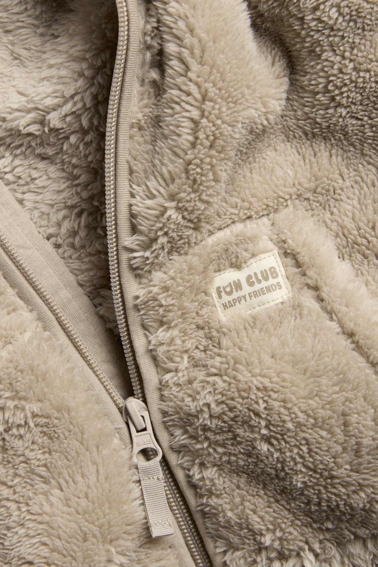 Zbliżenie beżowej kurtki polarowej z zamkiem błyskawicznym i metką "FUN CLUB" na piersi.