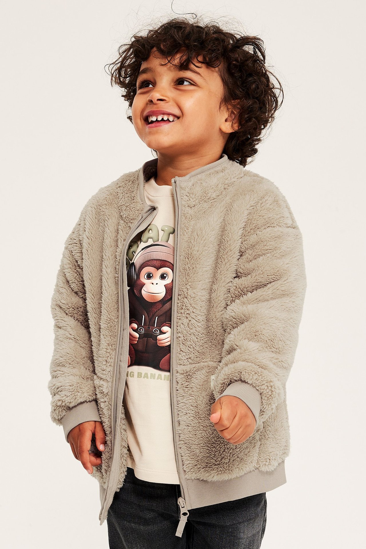 Etunäkymä: hymyilevä lapsi beigessä piletakissa, auki yllä apinaprinttipaidan ja tummien farkkujen päällä.