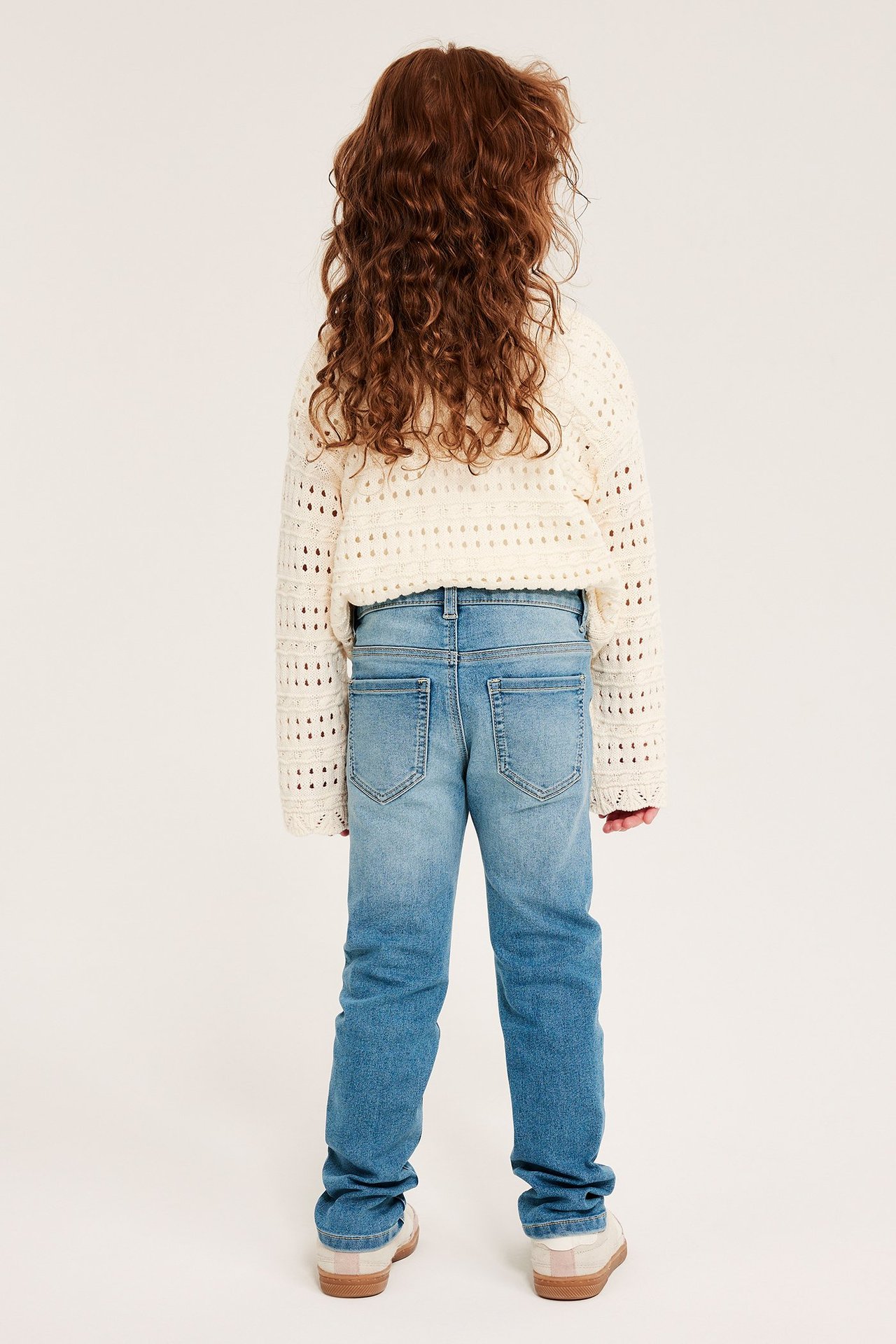 Widok z tyłu dziecka pokazuje jasnoniebieskie jegginsy slim, krótki kremowy sweter szydełkowy i jasne buty.