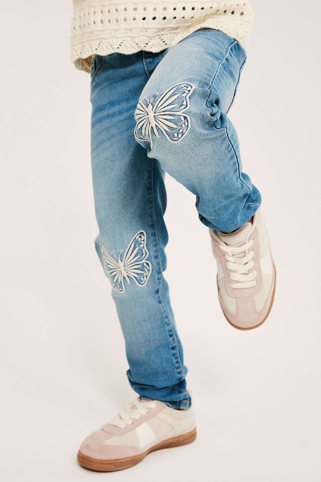 Widok z przodu dziecka w jasnoniebieskich legginsach jeansowych z haftem motyli i jasnoróżowych tenisówkach.