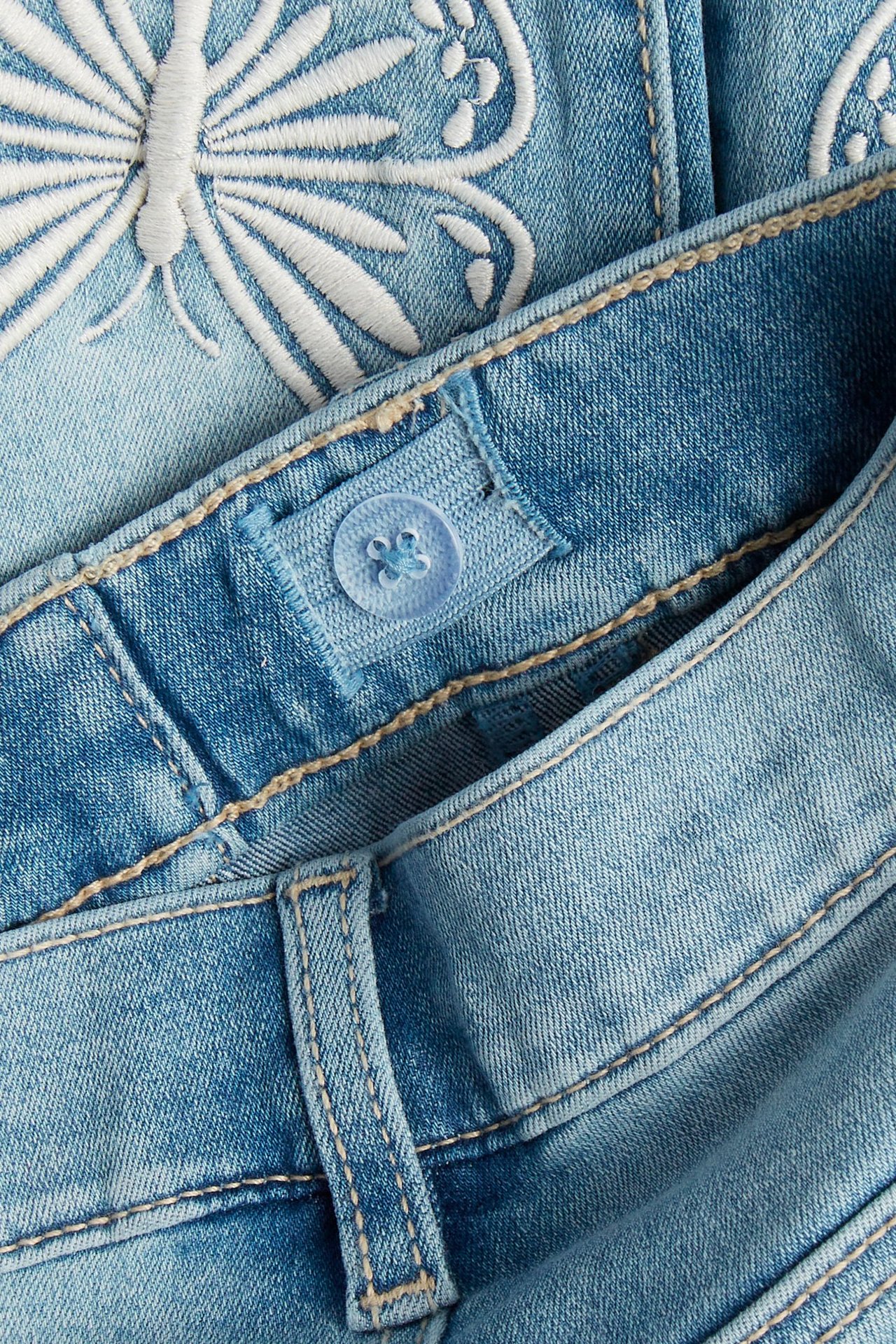 Zbliżenie na jasne dżinsowe jegginsy slim z białym haftem motyla i niebieskim guzikiem w talii.