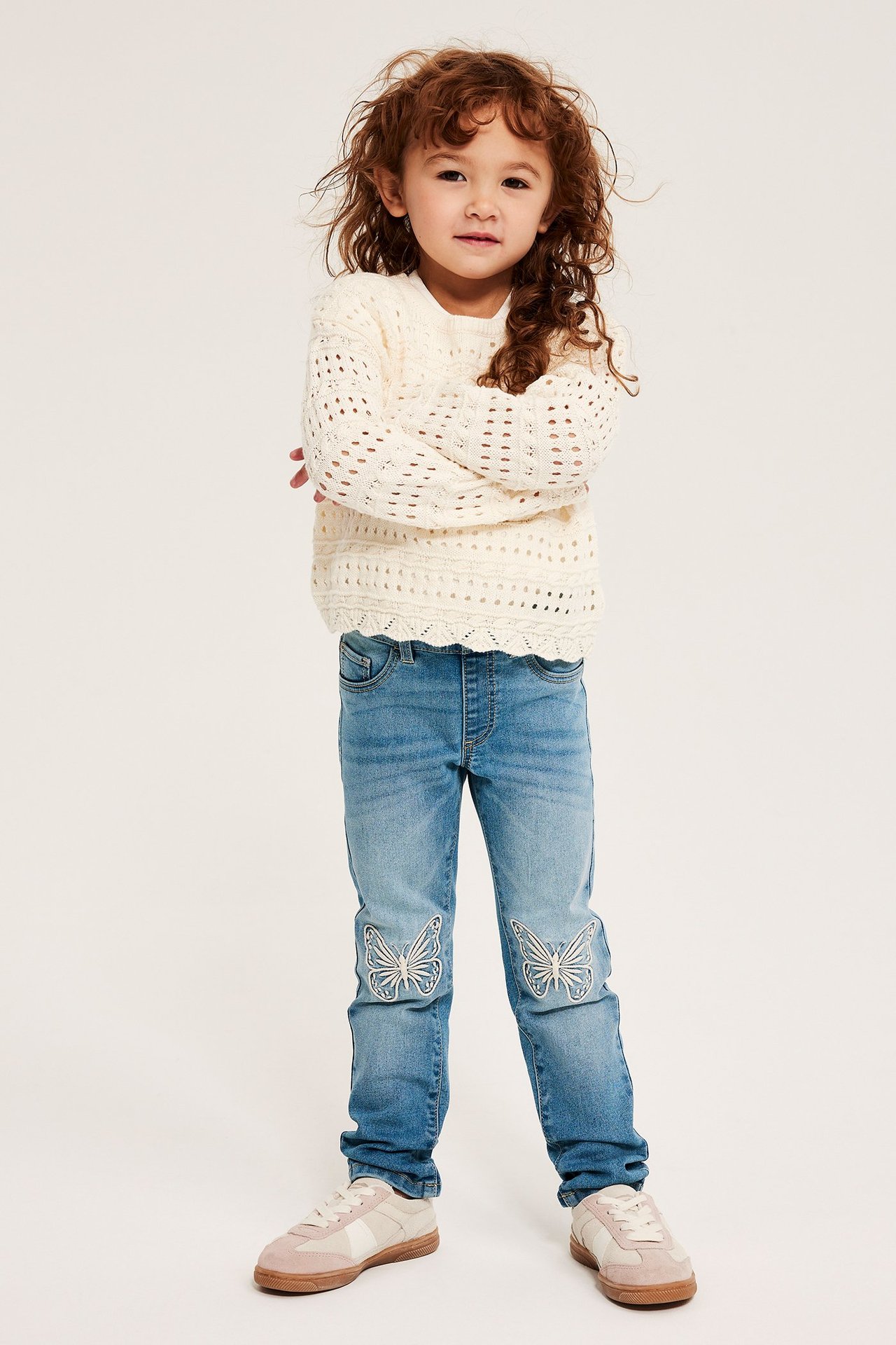 Widok z przodu dziecka w jasnoniebieskich legginsach dżinsowych z haftem motyli, kremowym swetrze i tenisówkach.