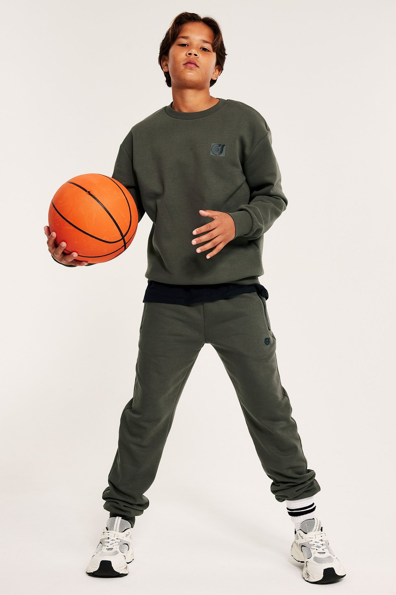 Chłopiec w ciemnozielonym dresie, trzyma piłkę do kosza, frontowe ujęcie.