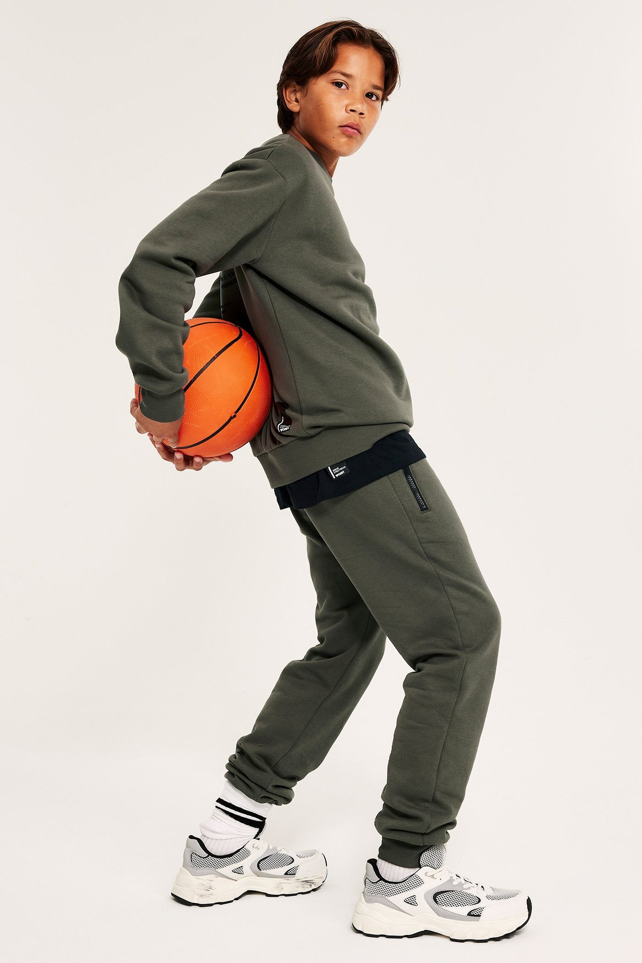 Chłopiec w zielonych spodniach dresowych, trzyma pomarańczowy basket, bok widok.
