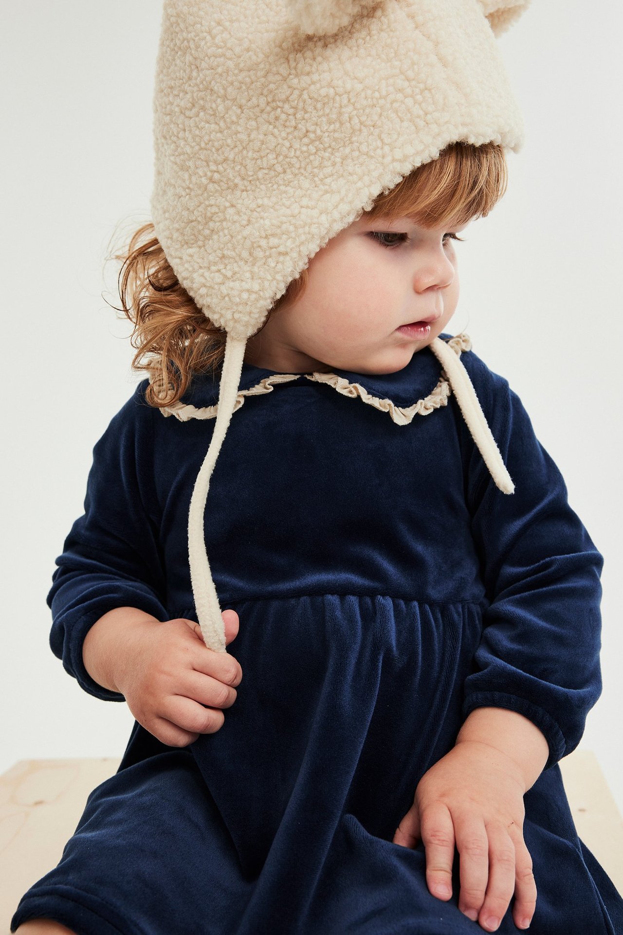 Närbild: baby i mörkblå velourklänning med volangkrage och gräddvit lurvig mössa, håller i bandet.