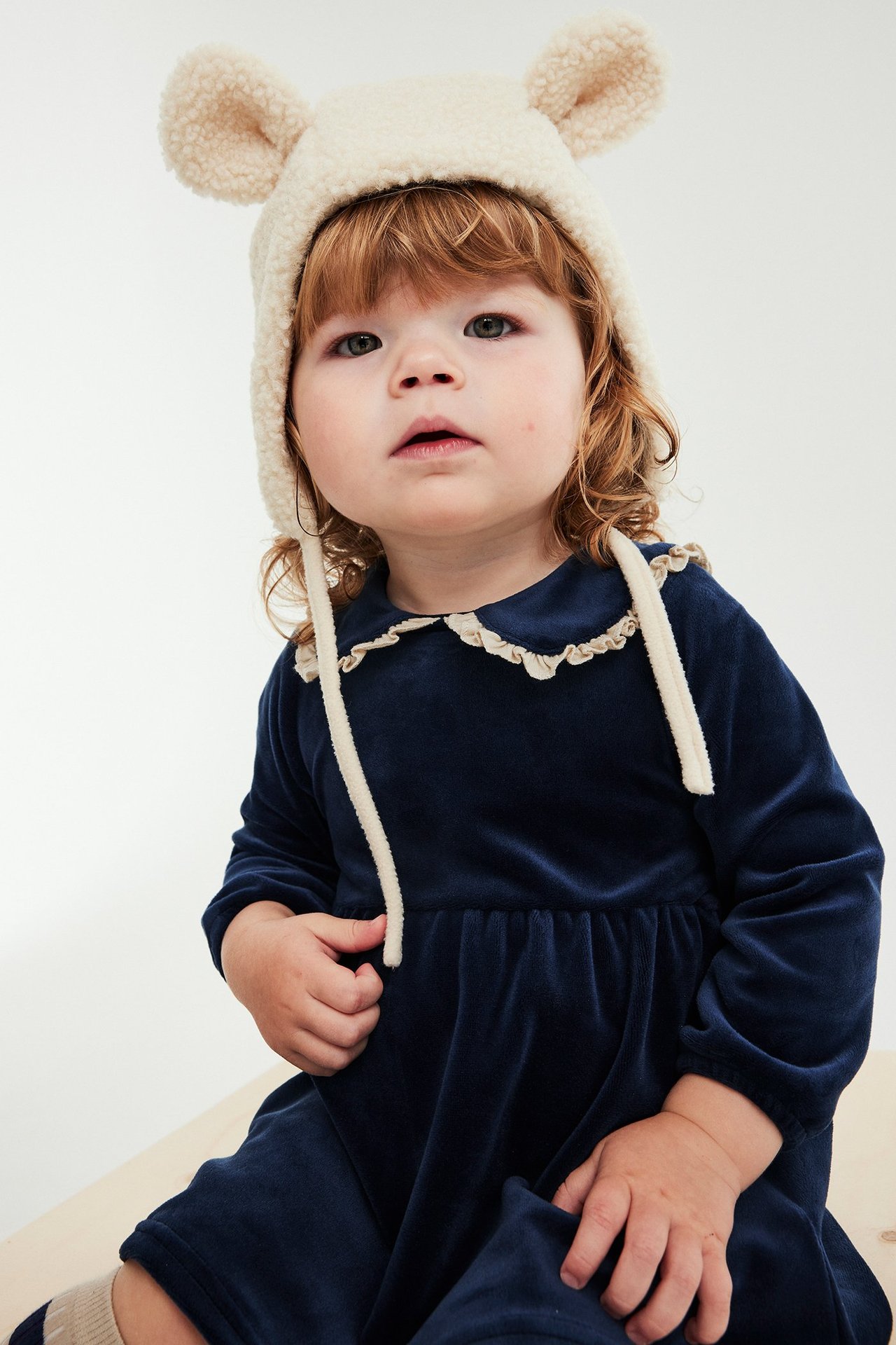 Nærbilde av baby i mørkeblå velourkjole med kremfarget volangkrage og kremfarget teddybjørnlue.