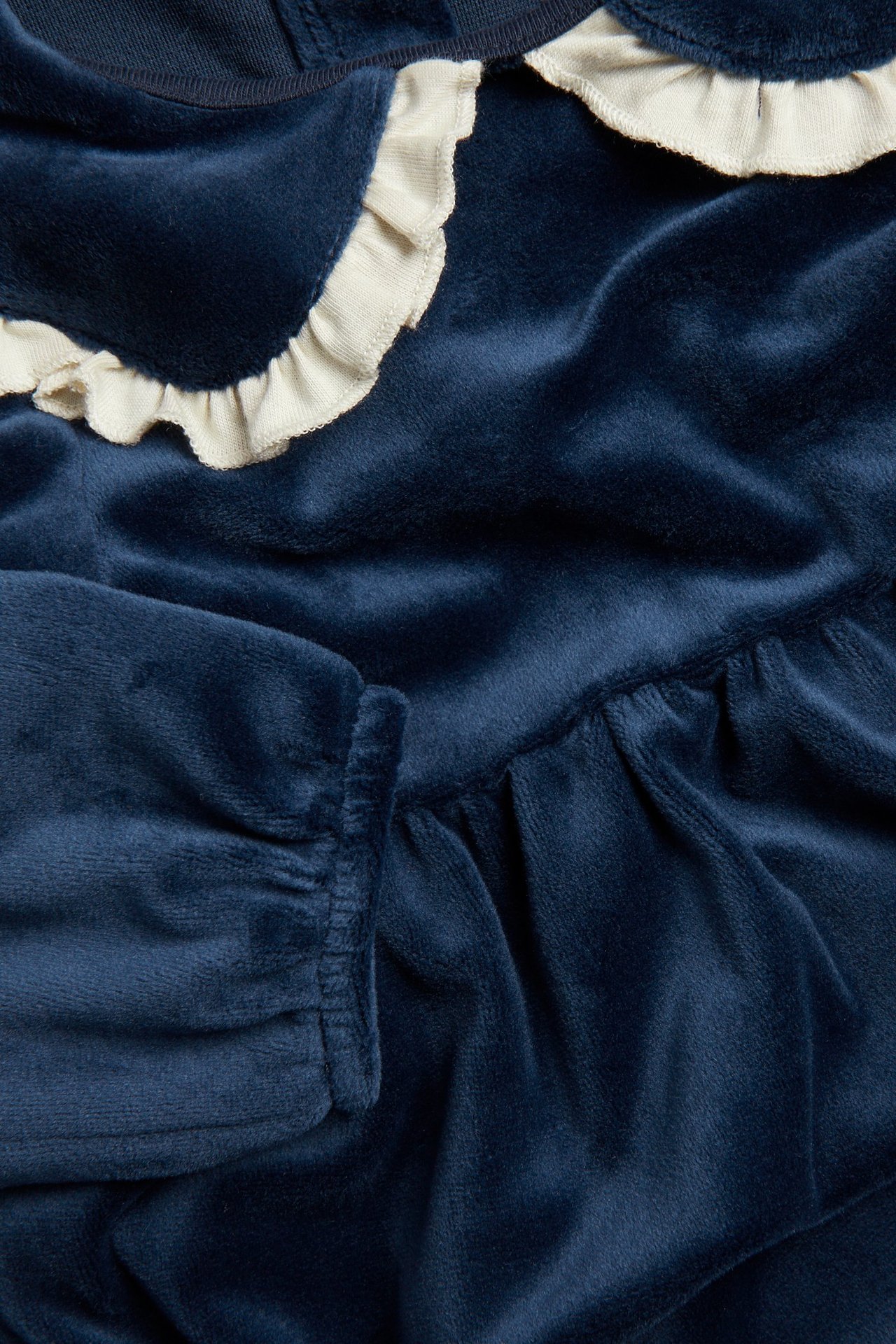 Zbliżenie granatowej welurowej sukienki niemowlęcej z kremowym falbankowym kołnierzem i marszczonymi mankietami.