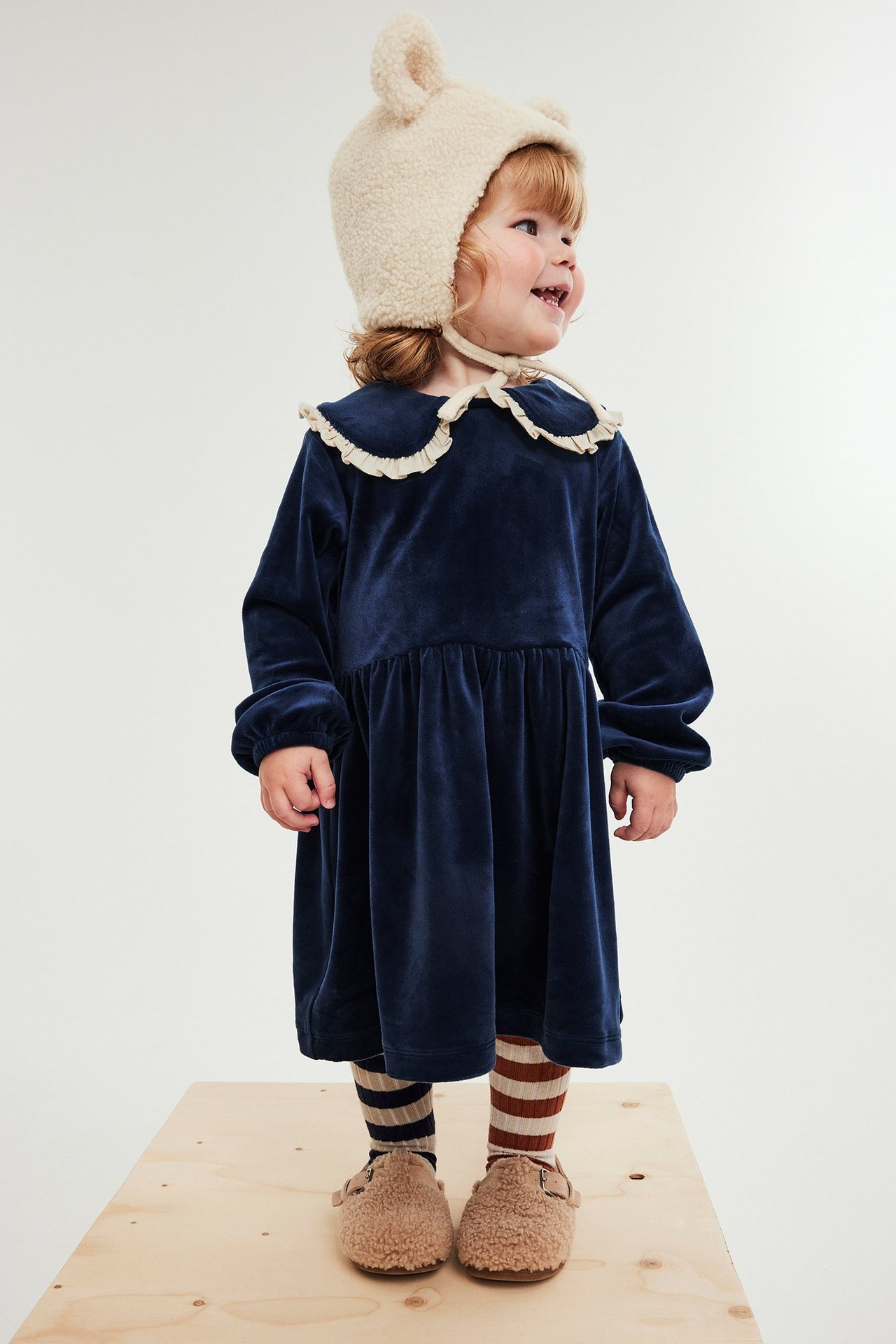 Widok z przodu: niemowlę w granatowej welurowej sukience z falbanką, pasiastych legginsach, futrzanych butach, czapce misia.